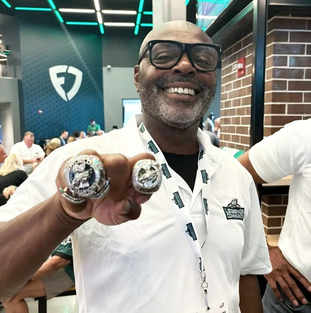 A smiling man wearing glasses and a white polo shirt with a 'Legends Community' logo, showing off large championship rings on his fingers at an indoor event, with a background of people and a blue and black wall.