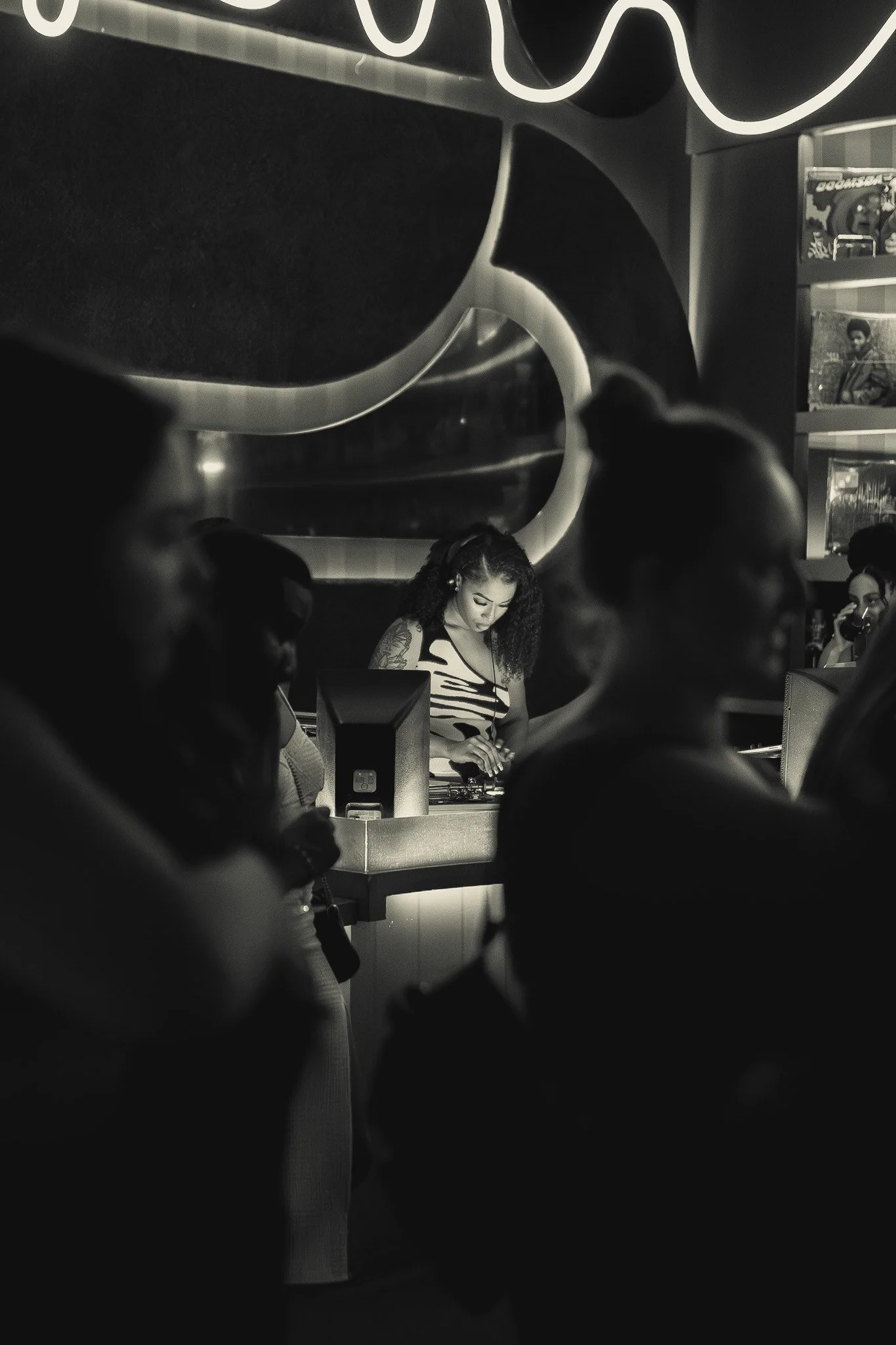A black and white photo of a woman DJ at a nightclub, with people in foreground and background, some with drinks, and a neon sign above.