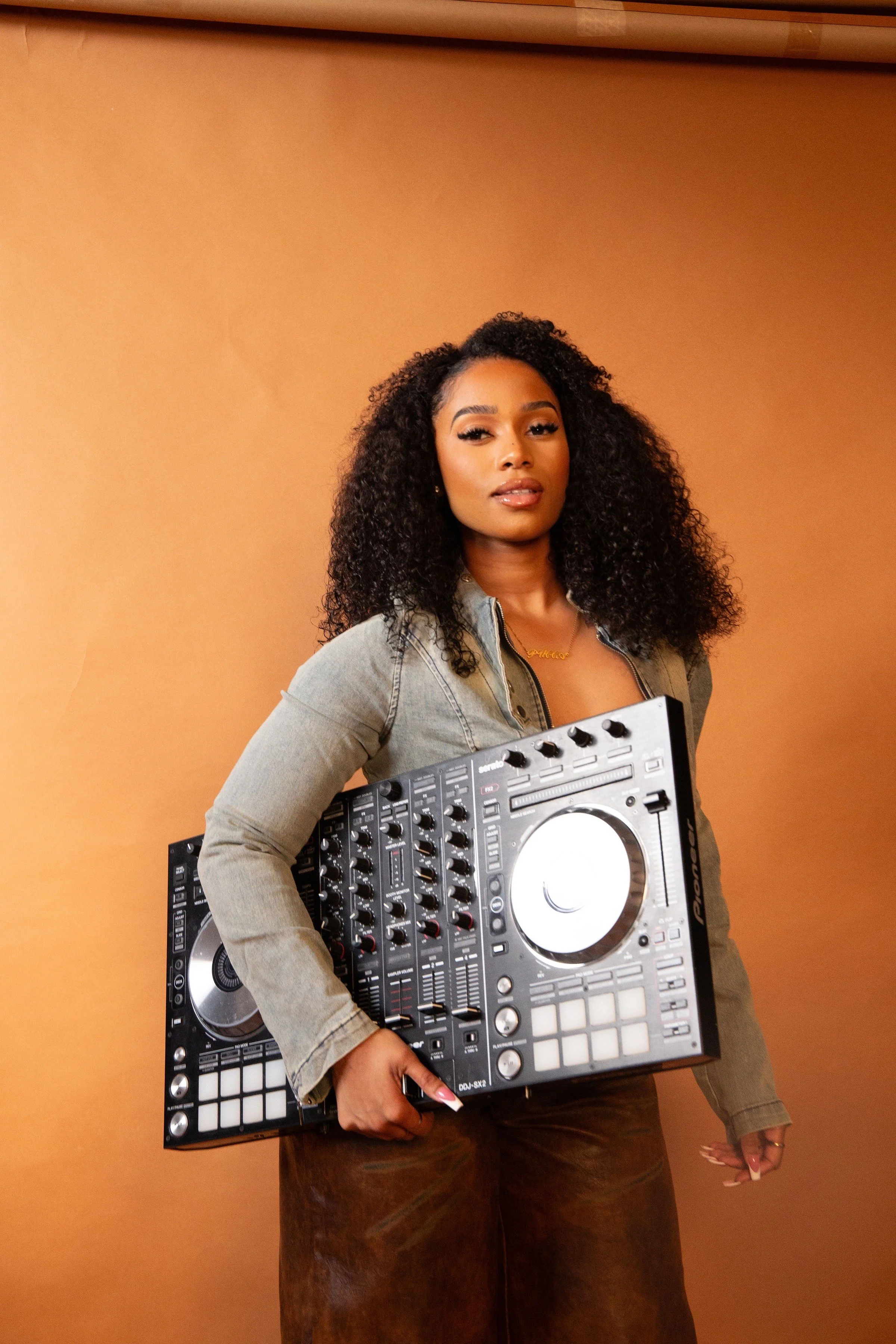 A Black woman with curly hair holding DJ equipment against a warm-colored background.