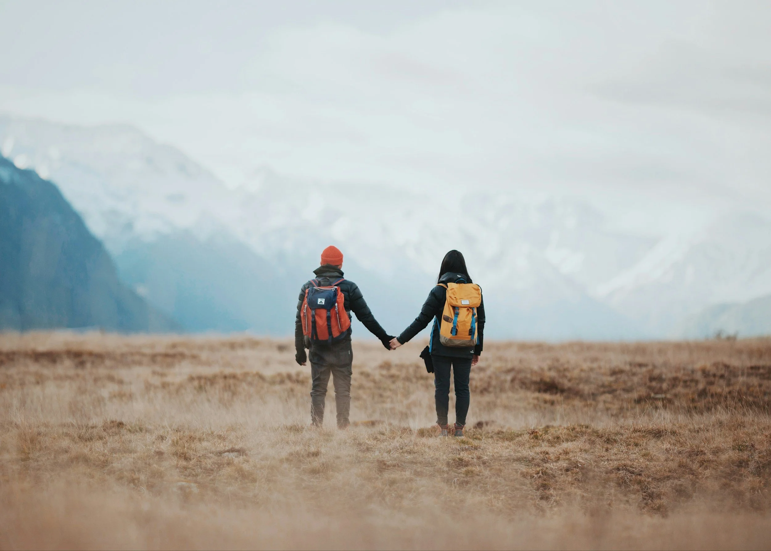 Two hikers, one man and one woman, hold hands while walking through a vast, open landscape with mountains in the background.