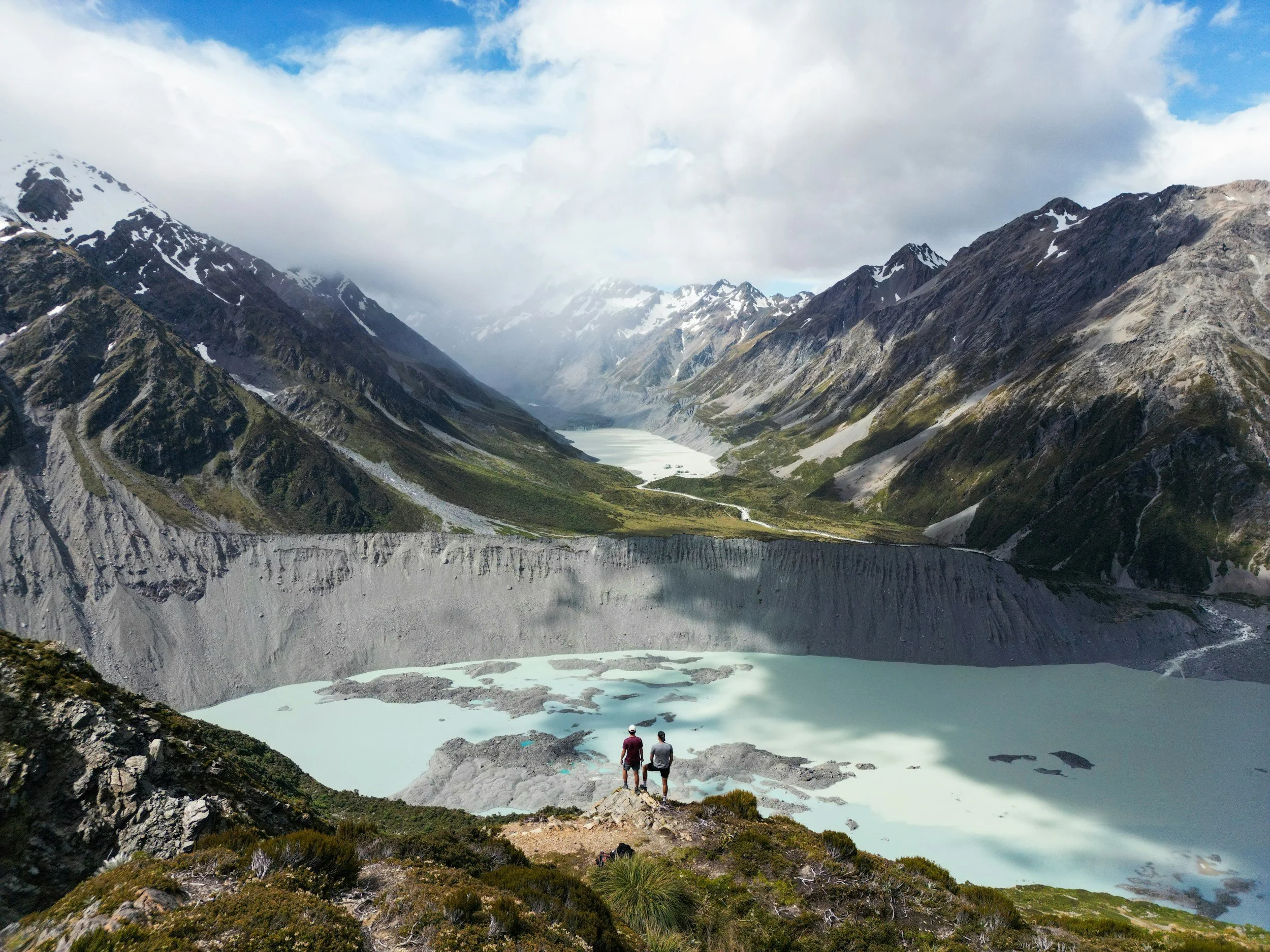Two hikers standing on a rocky ridge overlooking a glacial lake in a mountain valley with snow-capped peaks and clouds in the sky.