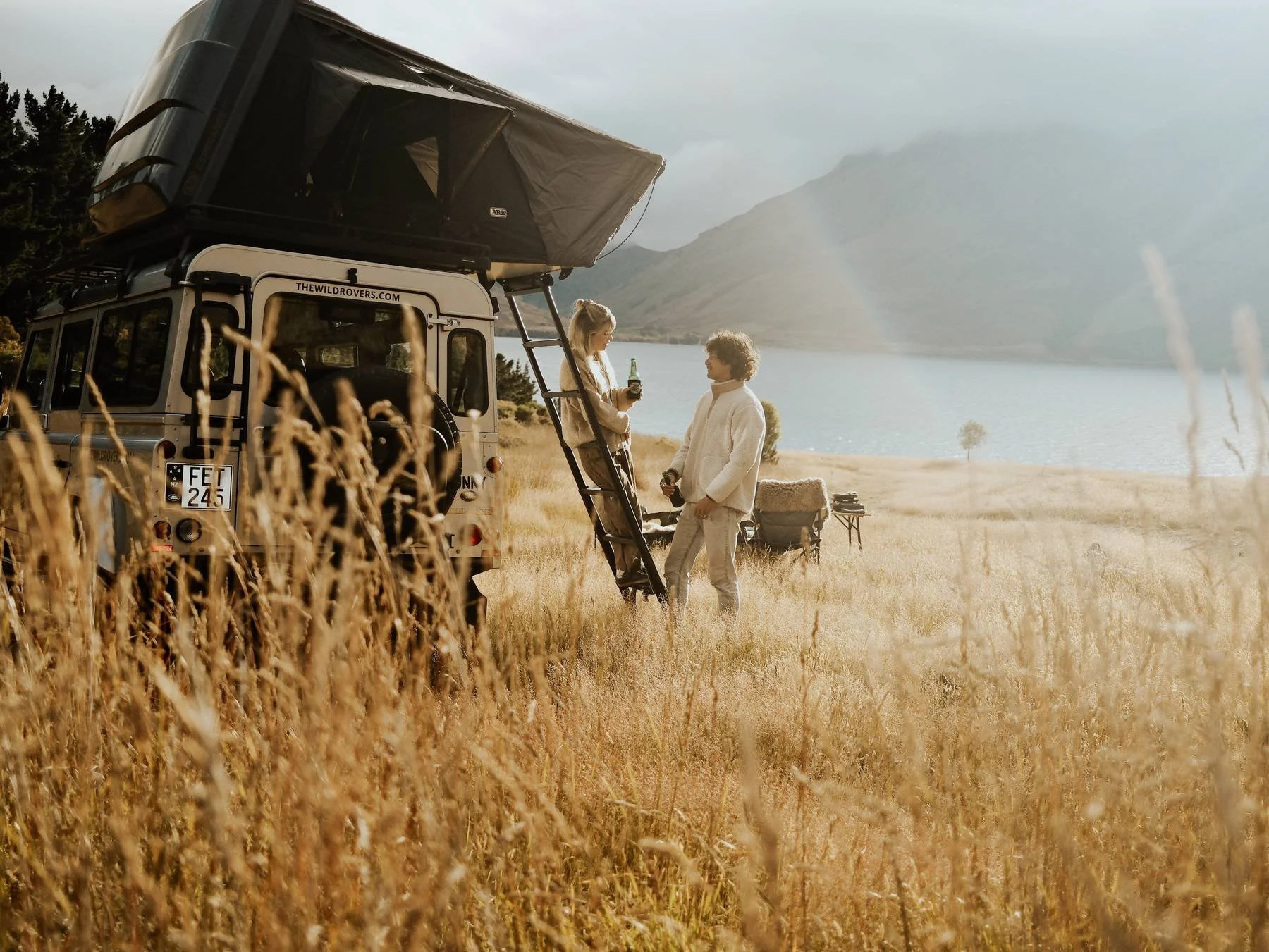 A couple standing near a camper van in a field of tall grass by a lake, with mountains and cloudy sky in the background.
