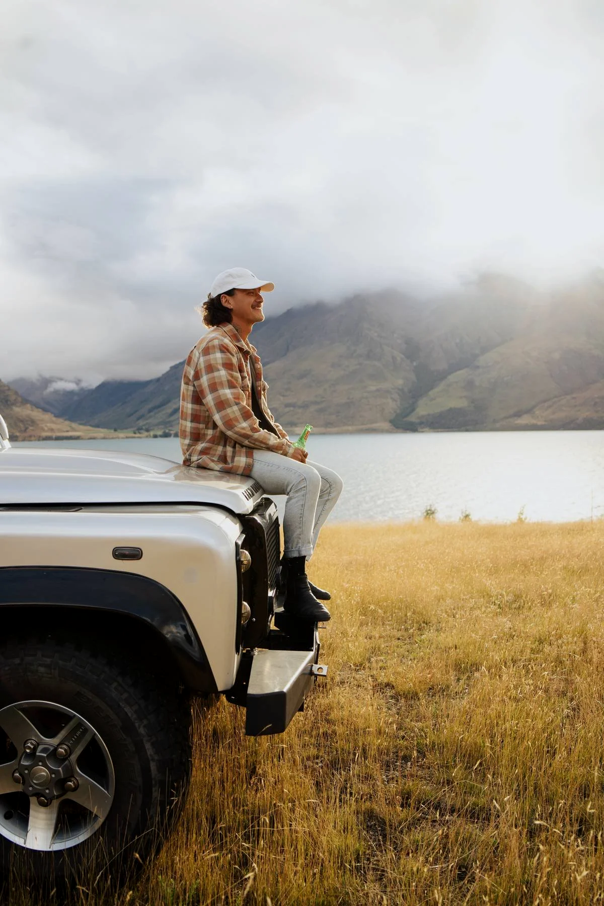 A person with curly hair, wearing a white cap and plaid shirt, sitting on the front of a white off-road vehicle, holding a drink, overlooking a lake and mountains under cloudy skies.