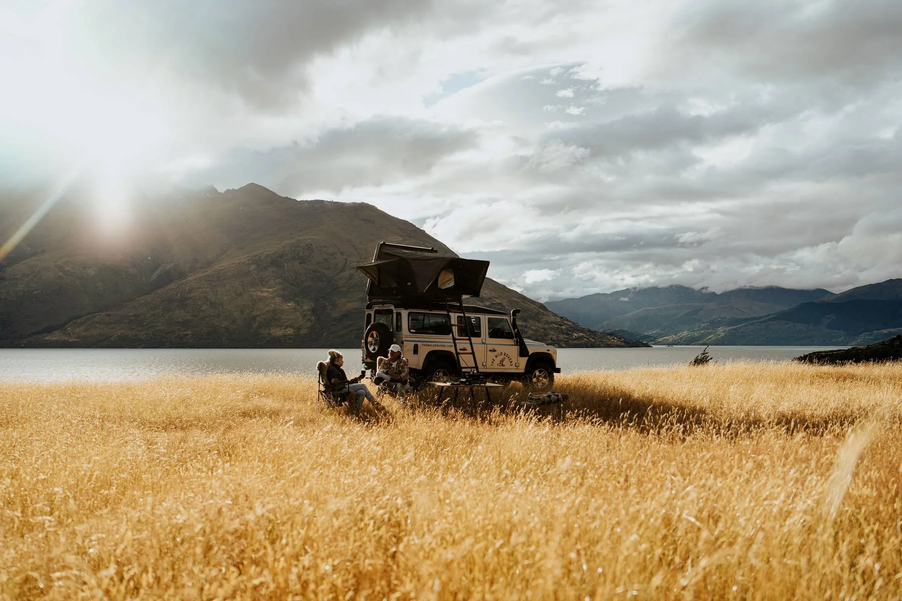 Two people sitting outdoors near a parked off-road vehicle in a grassy field with mountains, lake, and cloudy sky in the background.