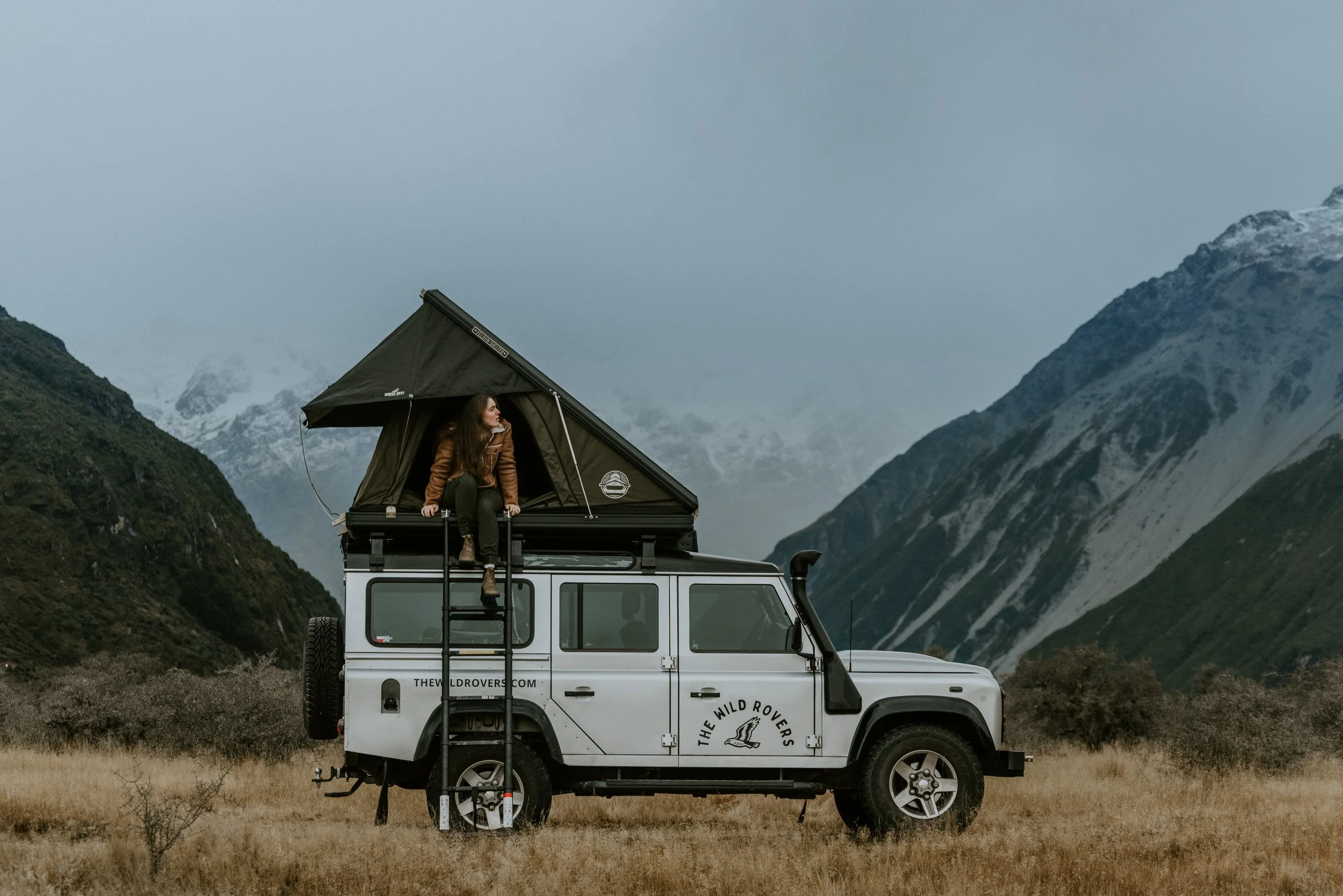 A woman sitting on top of a white Land Rover Defender in a mountainous landscape with snow-capped peaks and grassy fields, under an overcast sky.