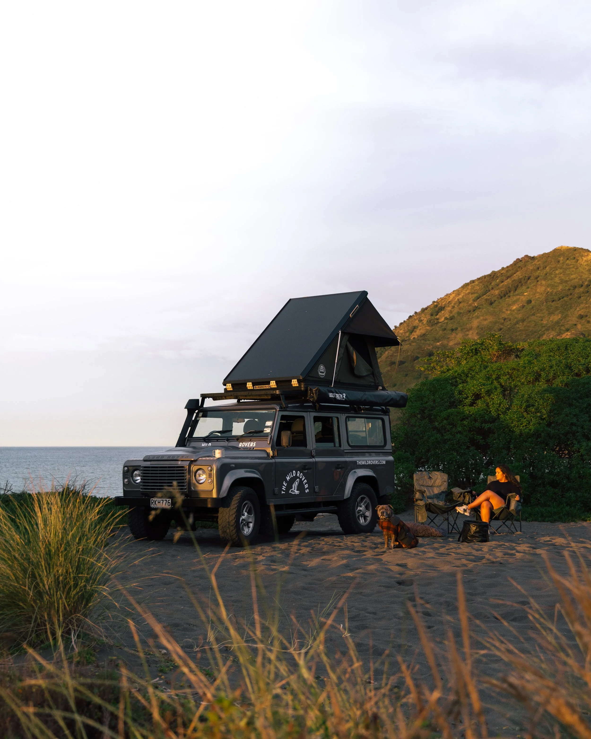 A black off-road vehicle with a rooftop tent parked on a beach, with a woman and a dog sitting on camping chairs nearby, and a hill with green vegetation in the background during sunset.