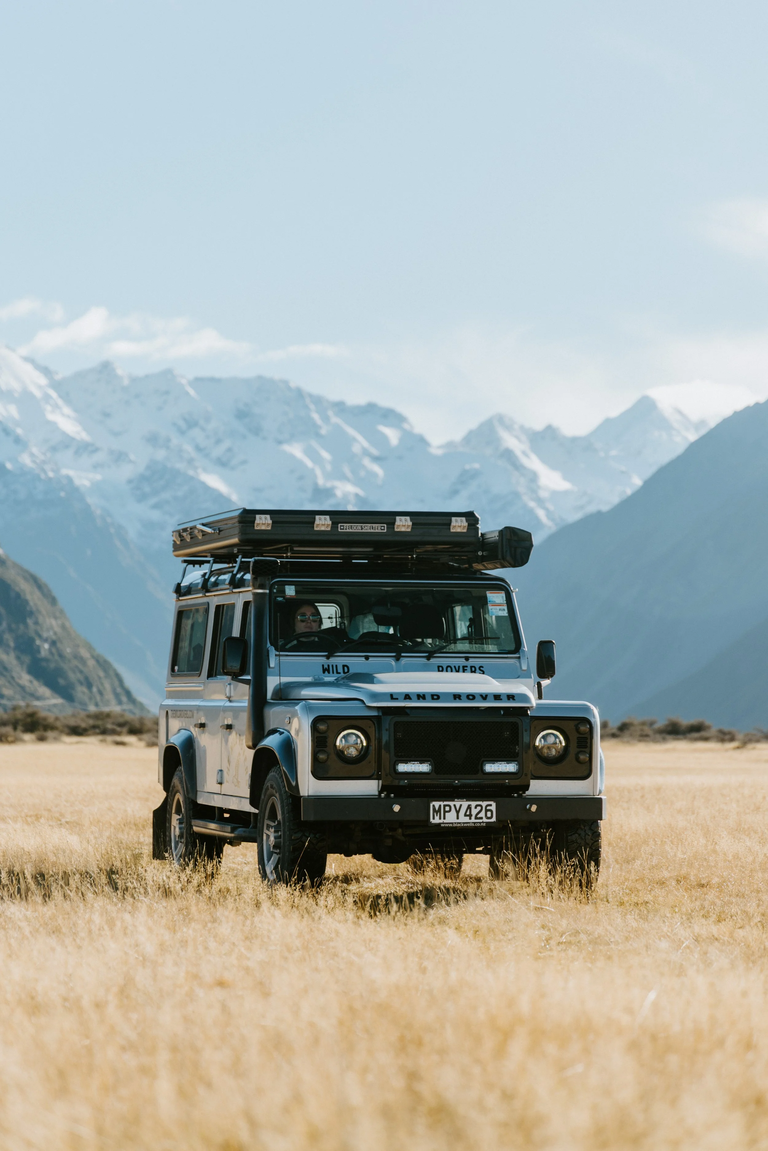 A white Land Rover Defender driving across a grassy plain with mountains in the background, under a cloudy sky.