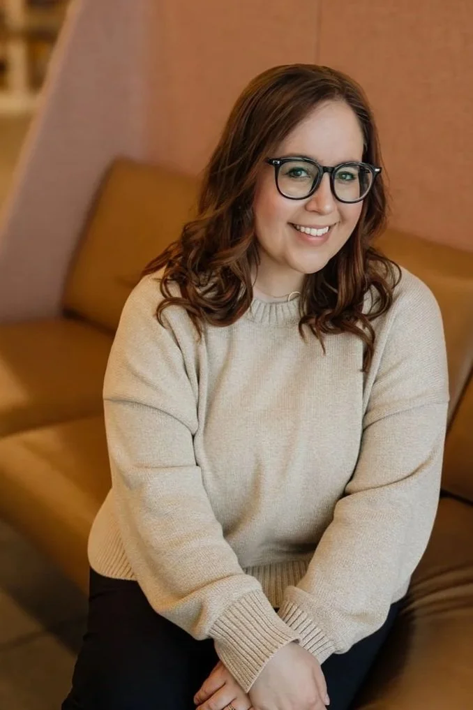 A young woman with brown wavy hair and glasses sitting on a couch, smiling at the camera, wearing a beige sweater.