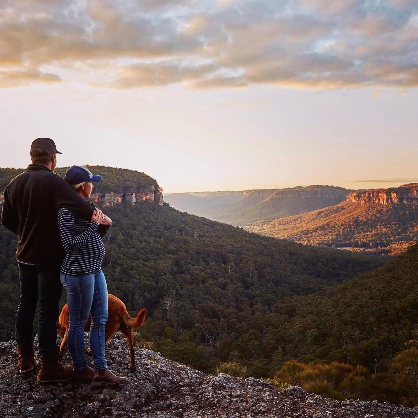 A couple with a dog overlooking a scenic valley at sunset, with rolling hills and cliffs in the background.
