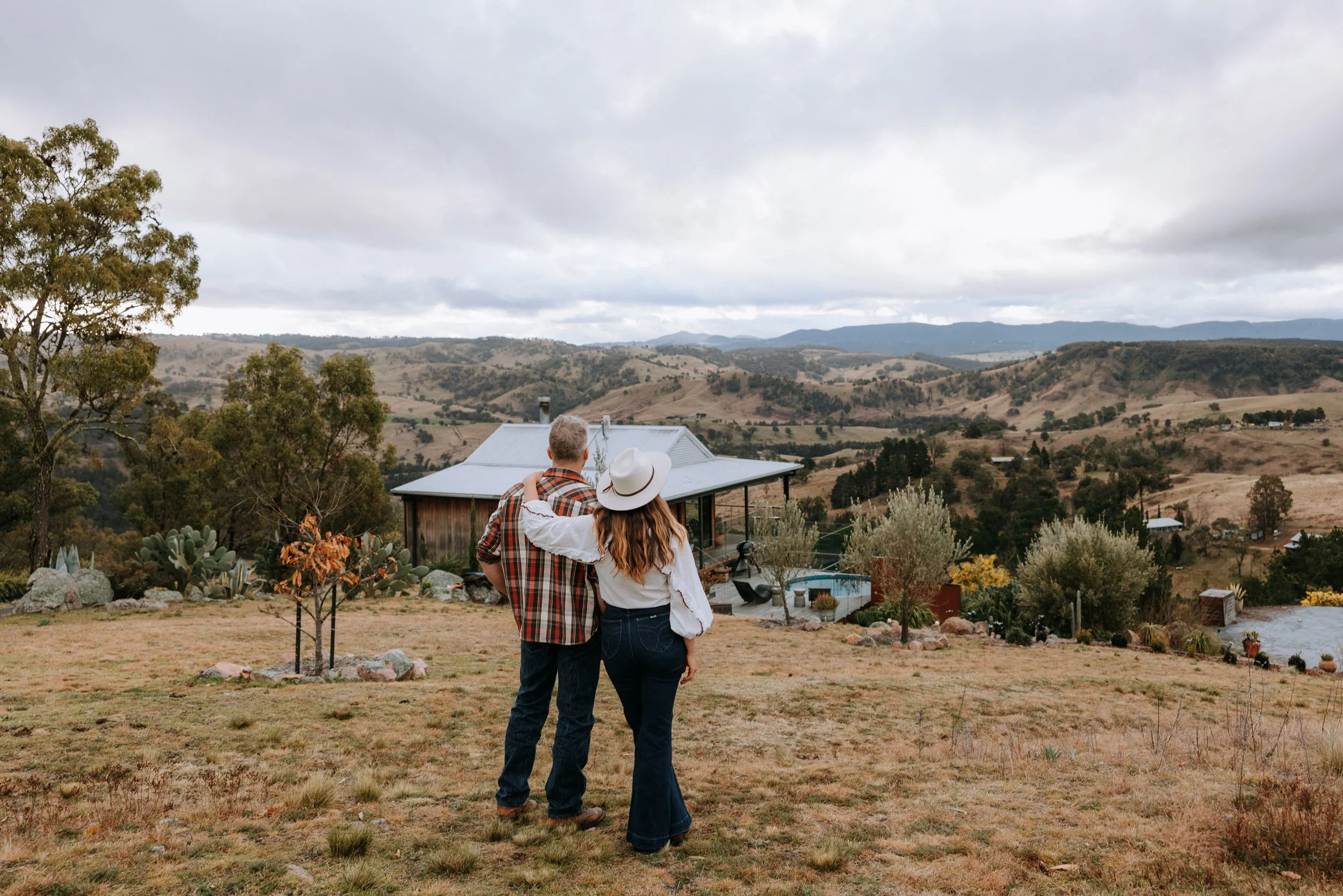 A couple standing close together outdoors, with a rural landscape and hills in the background, embracing and looking at the scenery.