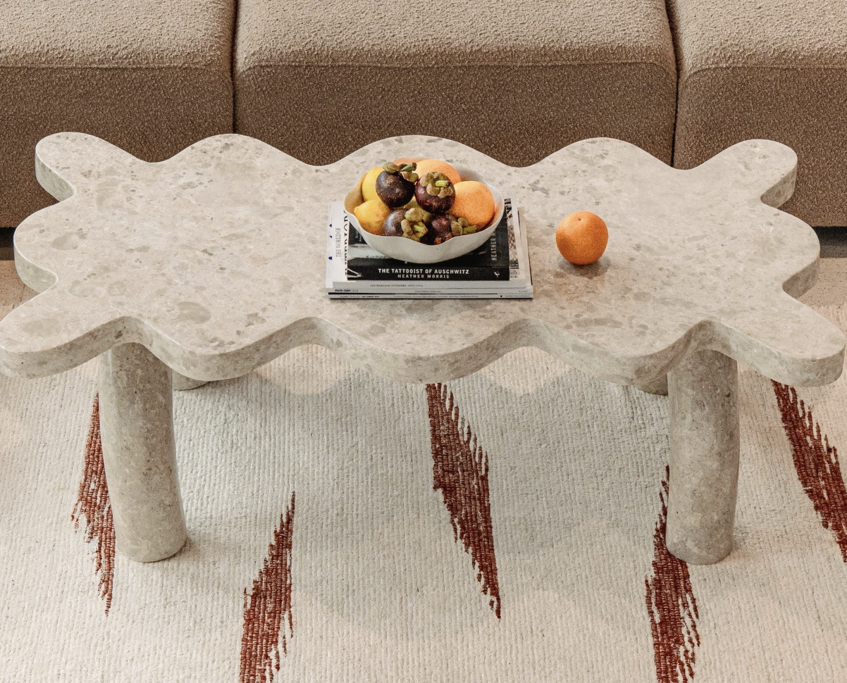 Contemporary beige stone coffee table with rounded edges and three cylindrical legs, placed on a cream-colored and red patterned rug. The table holds a white bowl of assorted fruit, a single orange, and a stack of magazines. Beige sofa is visible in the background.