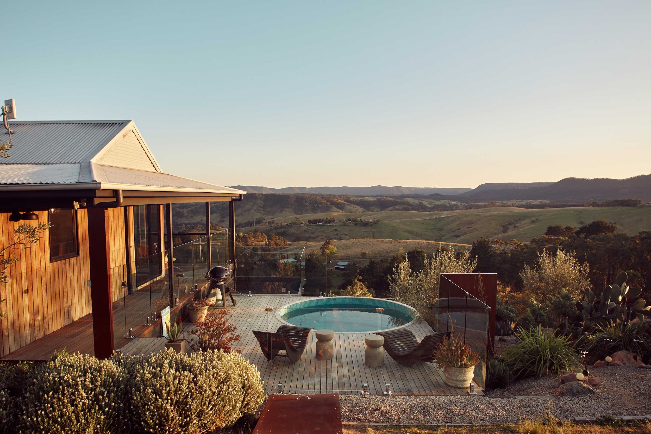 A balcony with a small round pool, two lounge chairs, and potted plants, overlooking rolling green hills at sunset.