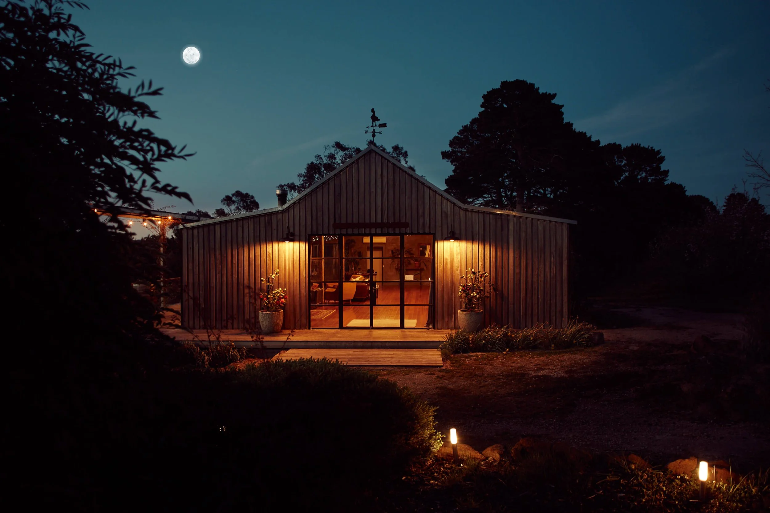 A cozy wooden house illuminated at night with a large full moon in the sky, surrounded by trees and outdoor lighting along a pathway.