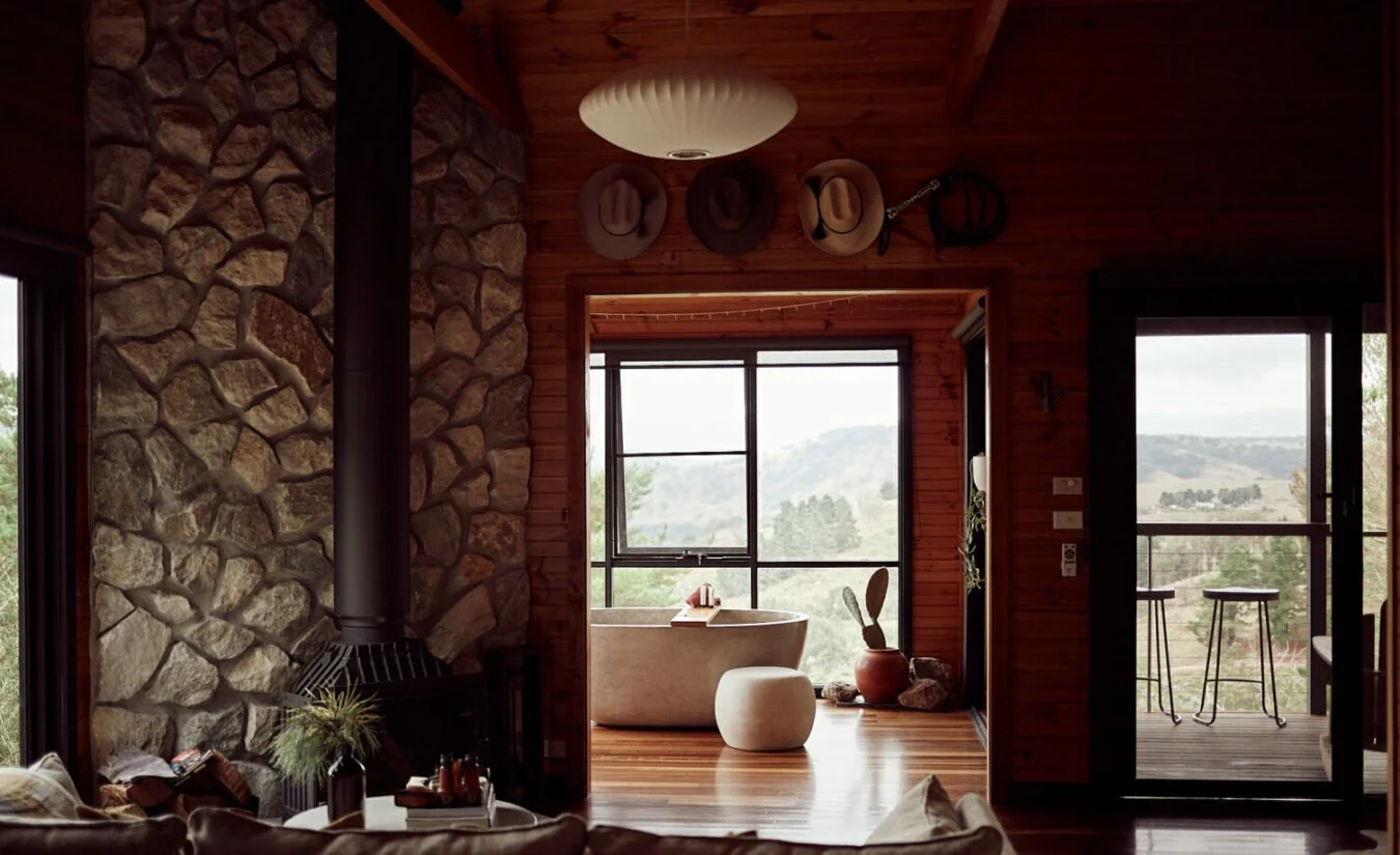 Interior of a wooden cabin with a stone fireplace, view of a bathtub in a sunroom, and a sliding glass door leading to a balcony with outdoor bar stools, overlooking a mountainous landscape.