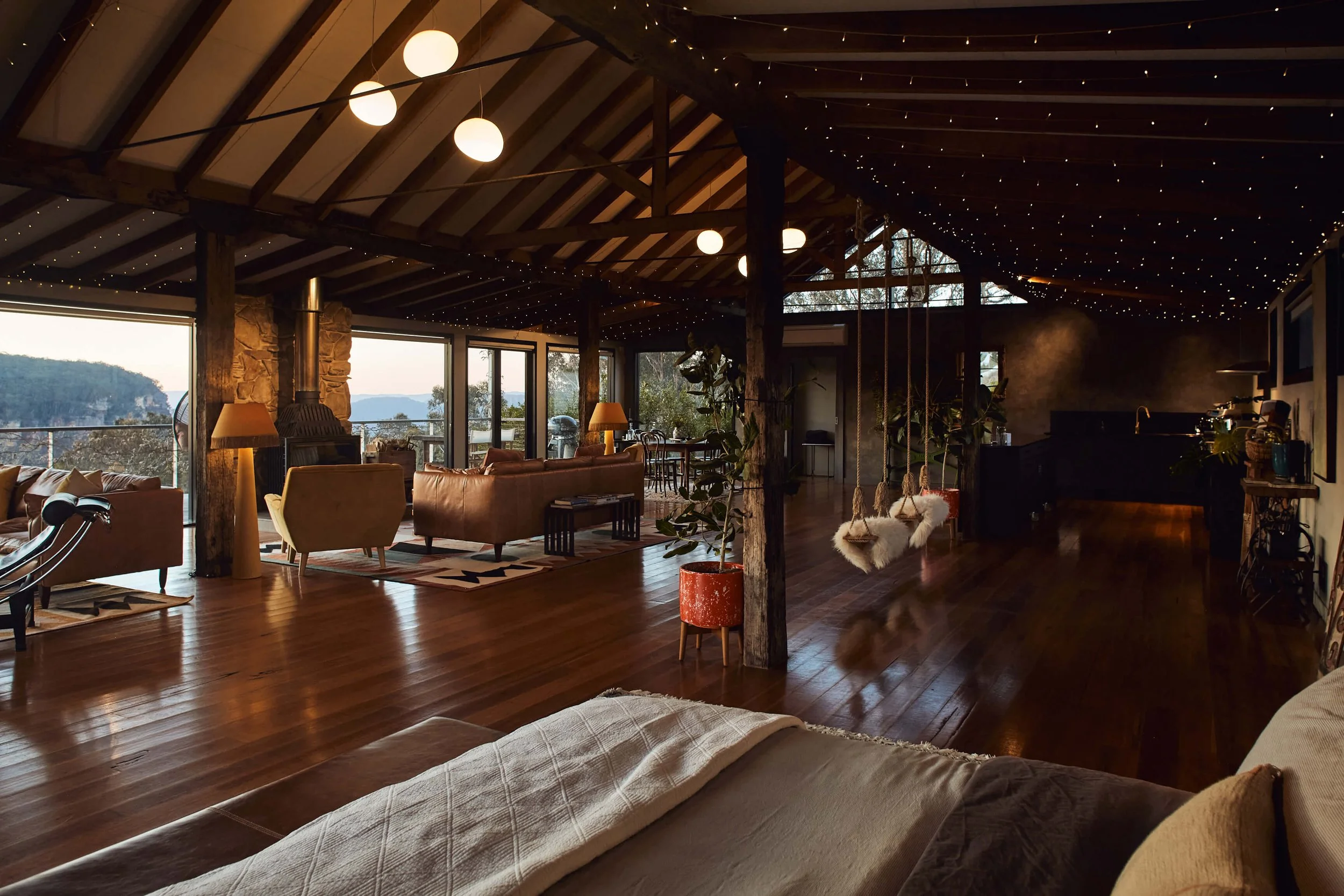 Spacious living room with wood floors, stylish furniture, string lights on ceiling, large window and glass doors revealing a scenic view, and a cozy ambiance.