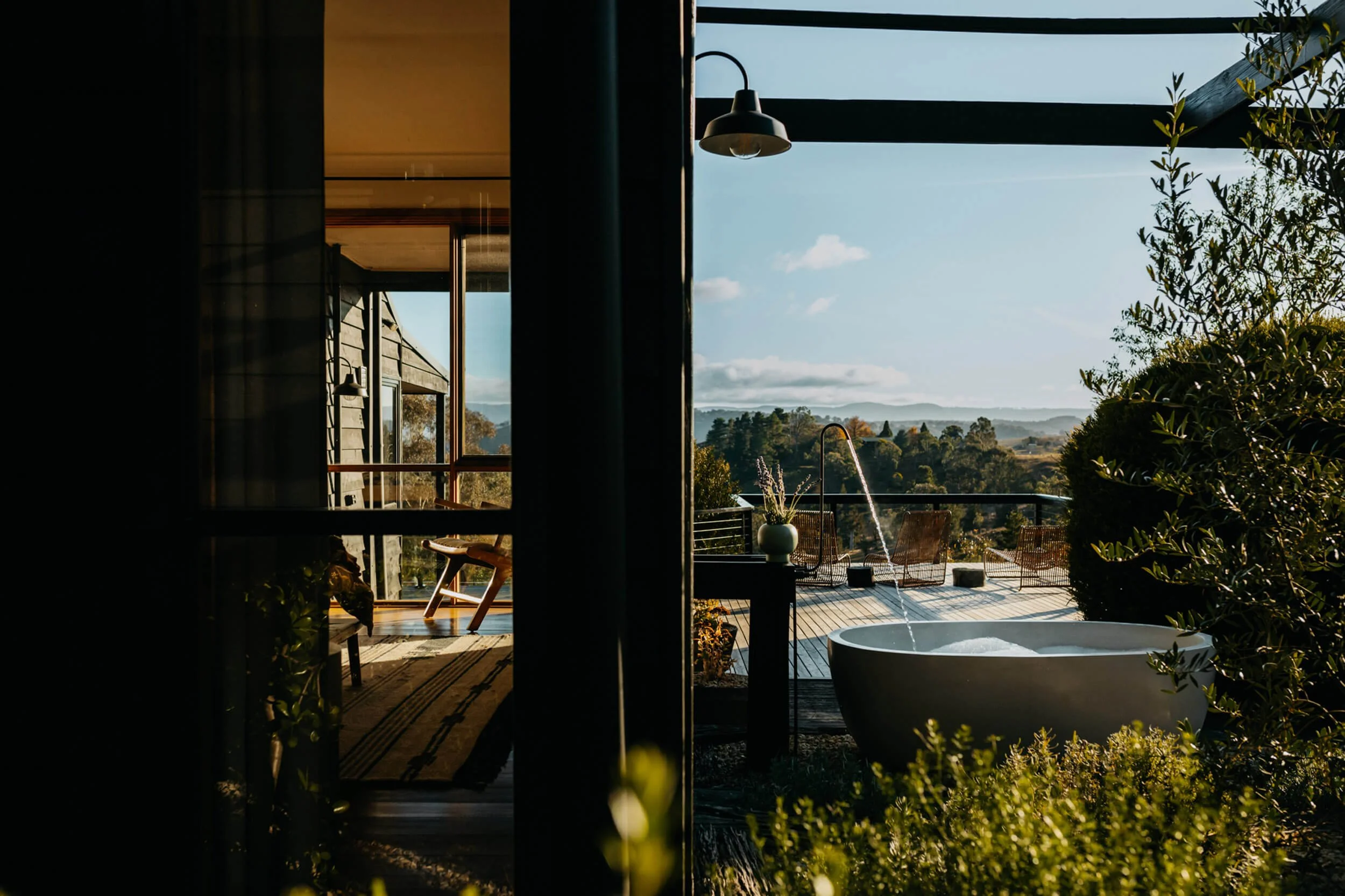 A scenic outdoor terrace with a large, round bathtub and lounge chairs, overlooking a lush landscape with trees and distant mountains under a clear sky.