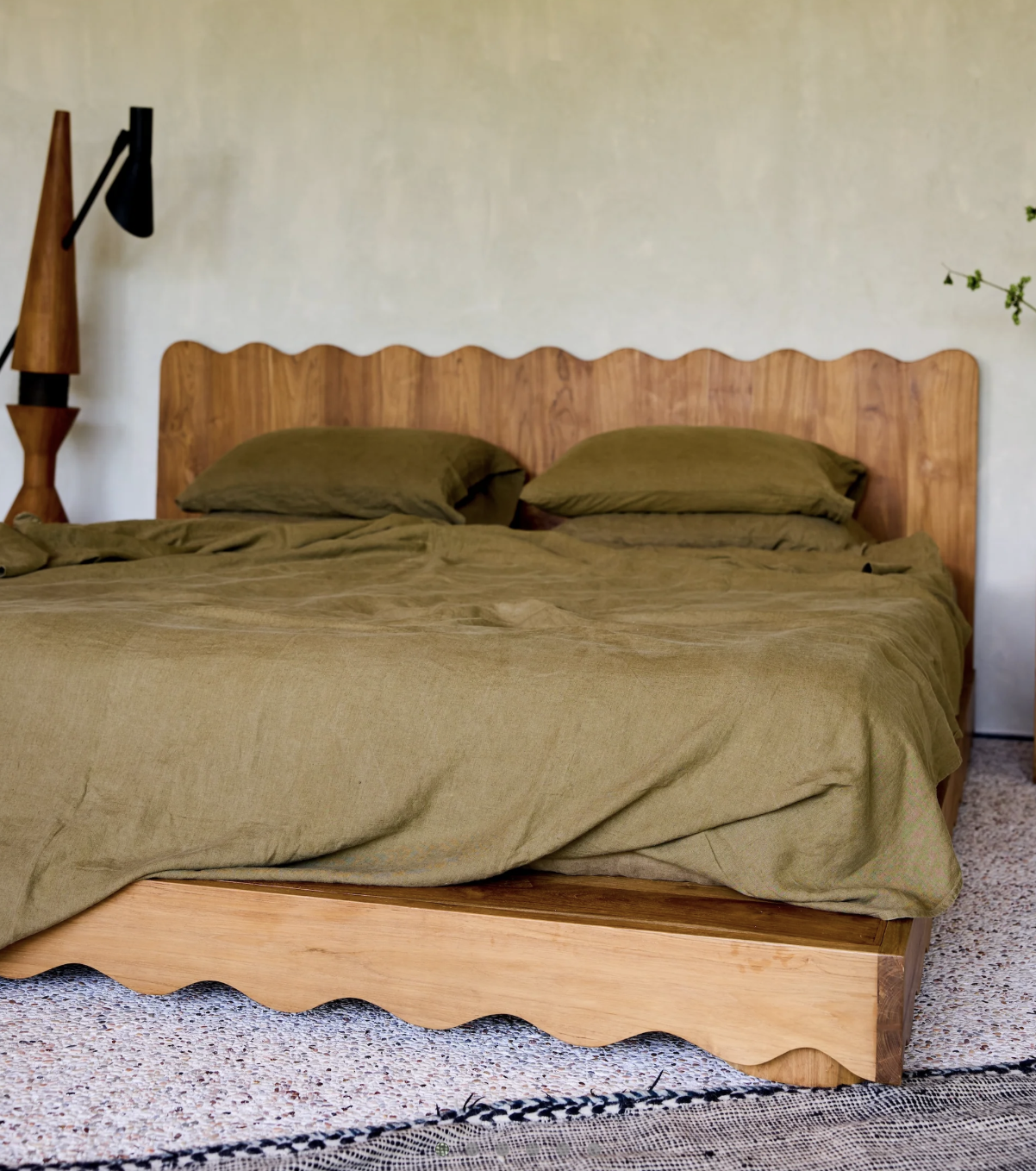 A wooden bed with a high scalloped headboard and olive green bedding and pillows, situated in a bedroom with a white wall and a patterned carpet.