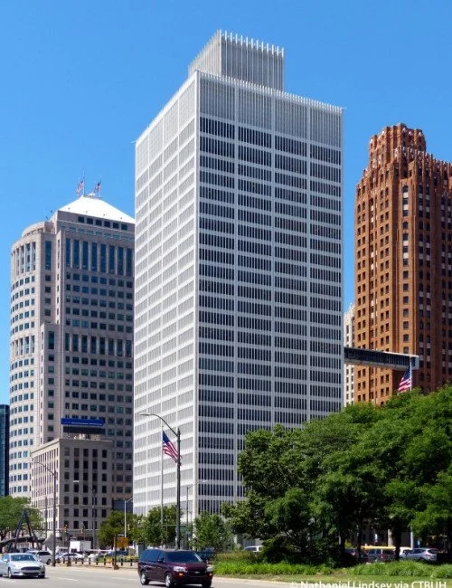 A cityscape of tall office buildings with a clear blue sky. In the foreground, there is greenery and a street with moving cars and American flags.
