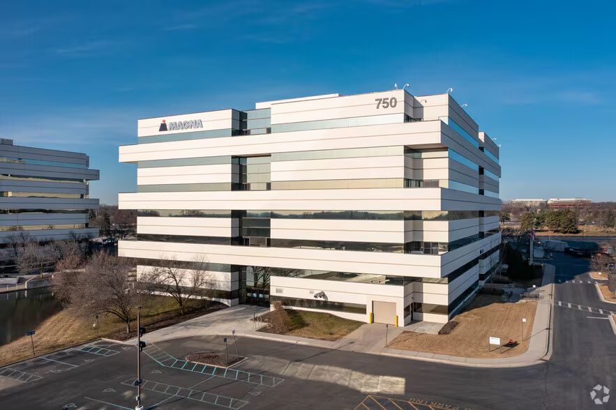 Modern white office building with black window accents and the MAGNA logo on the top left corner, located at 750. Clear sky and parking lot in front.