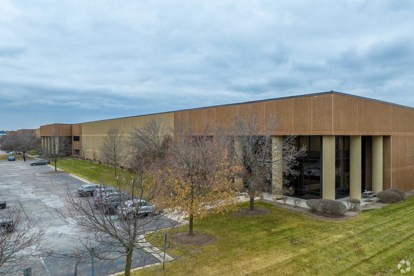 Large commercial building with brown and beige exterior, tall glass windows, and multiple columns at entrance, surrounded by leafless trees and parking lot, under a cloudy sky.