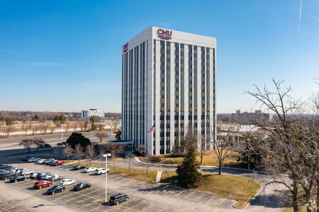A tall office building with the logo CMU for Central Methodist University on the top, surrounded by a parking lot with cars and leafless trees, under a clear blue sky.