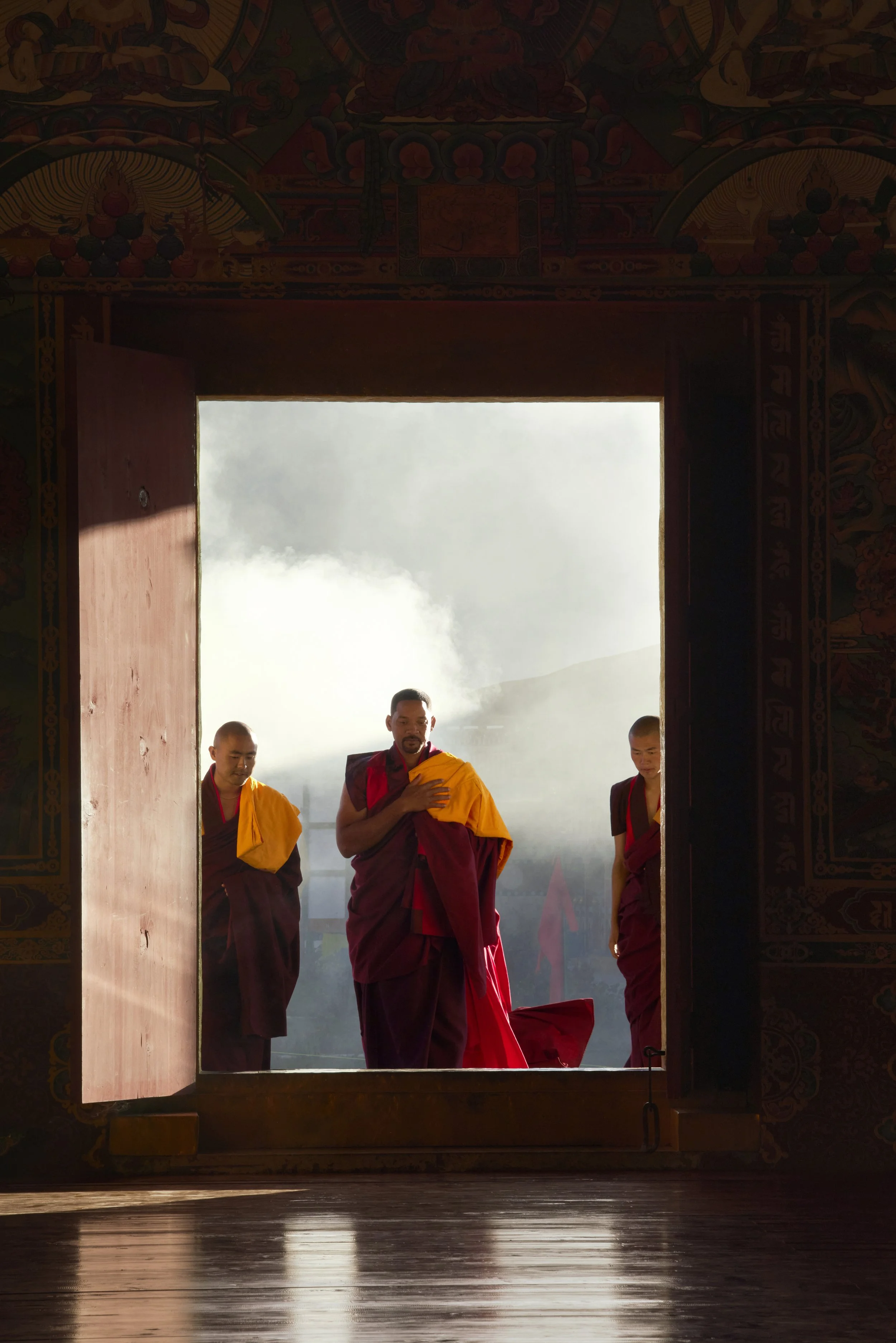 Will Smith and monks standing in the doorway of a temple, with mountains and mist in the background.