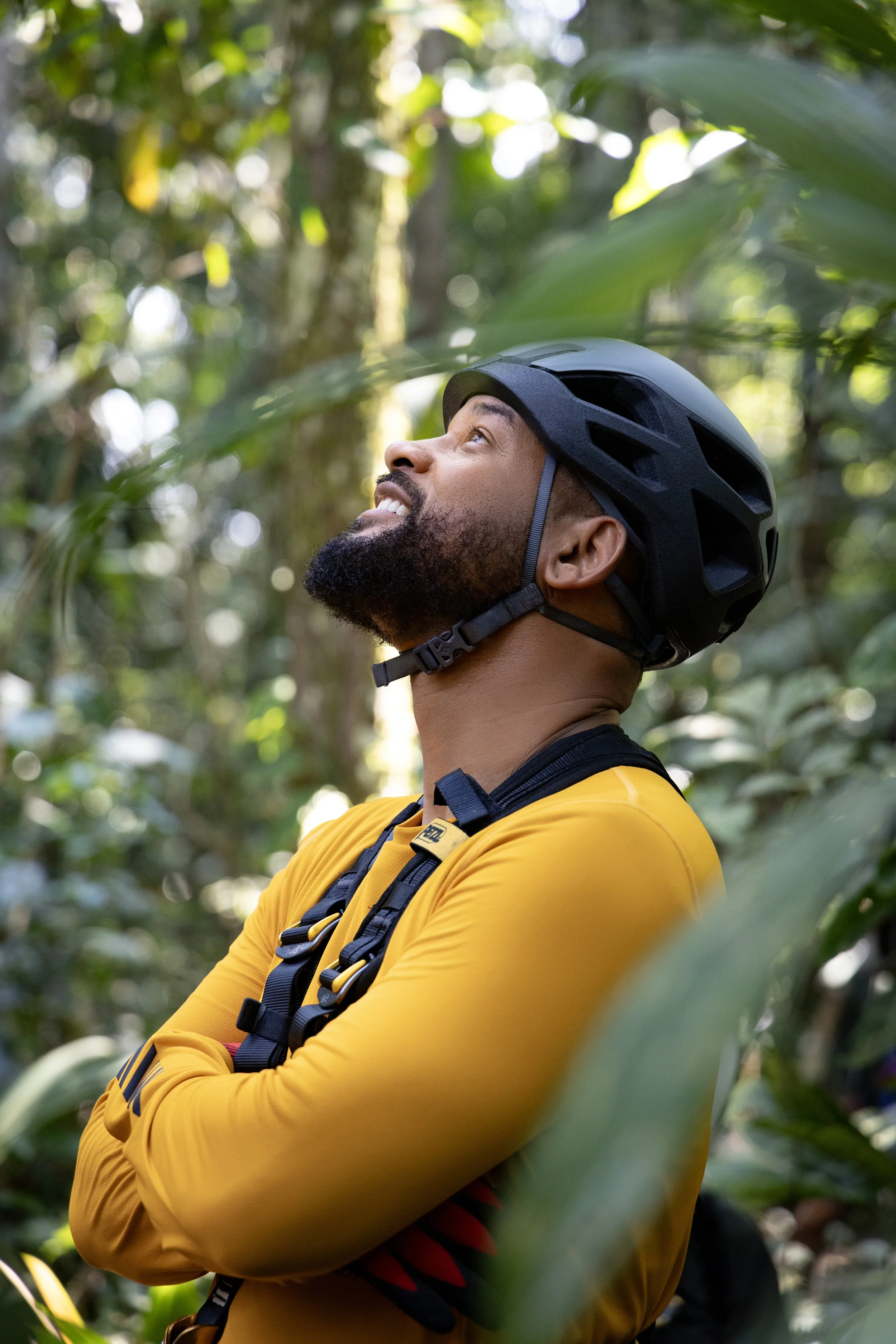 Will Smith wearing a black bike helmet and yellow long-sleeve shirt, standing in a forest, smiling and looking upward amidst green foliage.
