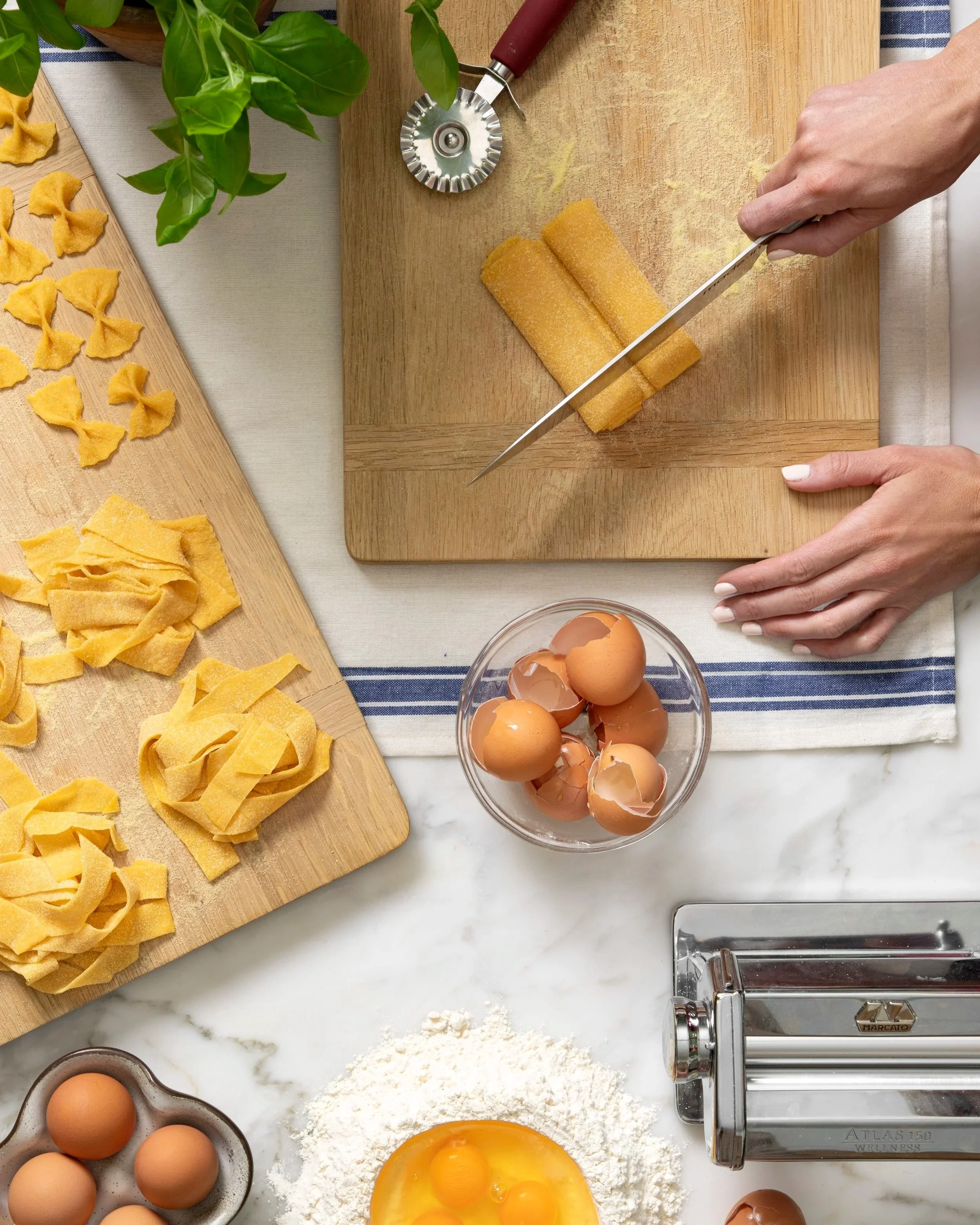 A person's hands are slicing uncooked pasta on a wooden cutting board. The scene includes eggs, pasta, flour, and a pasta machine on a white countertop with a striped cloth underneath.