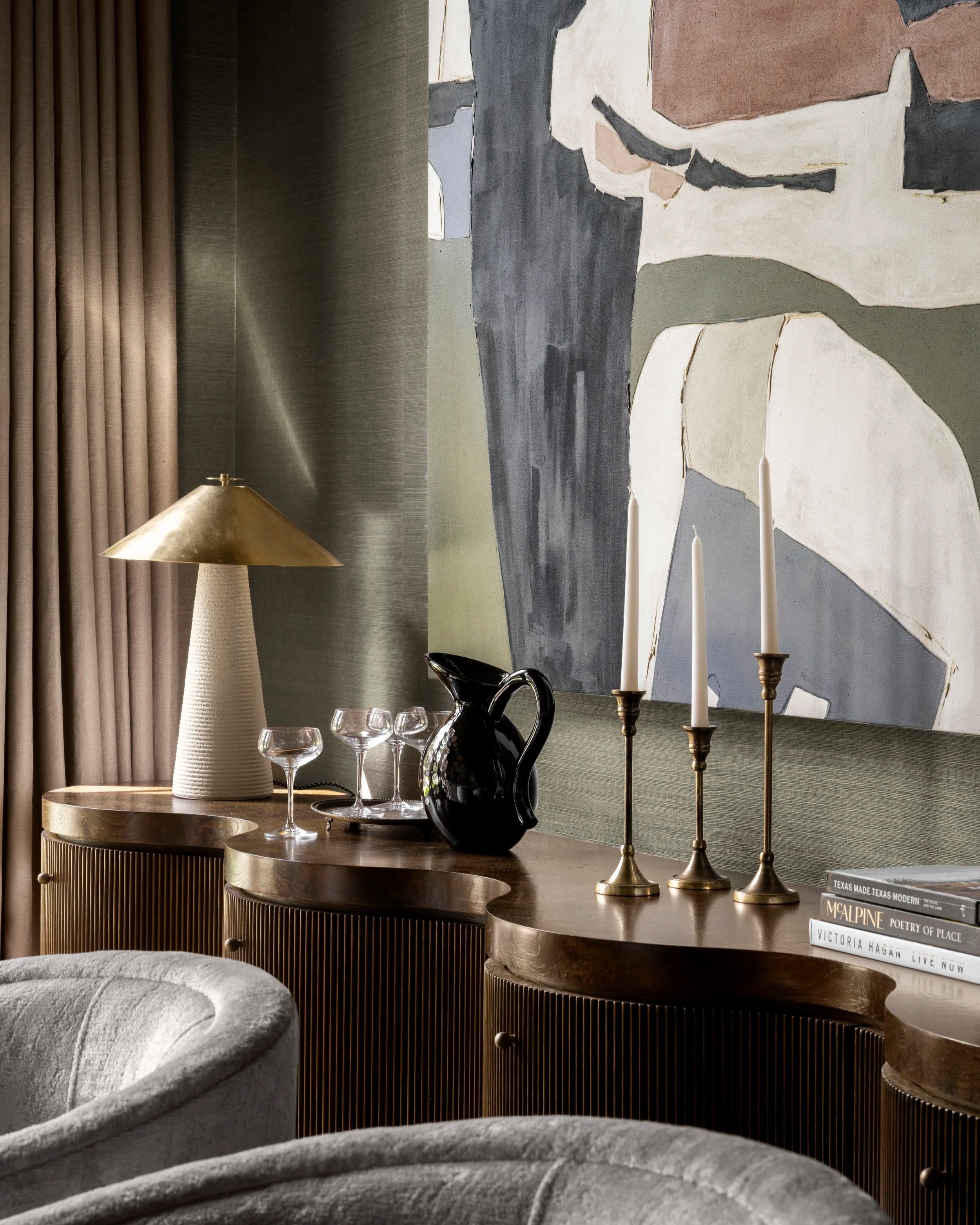 Interior scene with a curved wooden credenza displaying decorative items including a black vase, three white candles in gold holders, a brass lamp, and a glass cup. Behind, a textured wall with vertical tan curtains and abstract artwork with organic shapes and muted colors. Several books are stacked on the right side of the credenza.