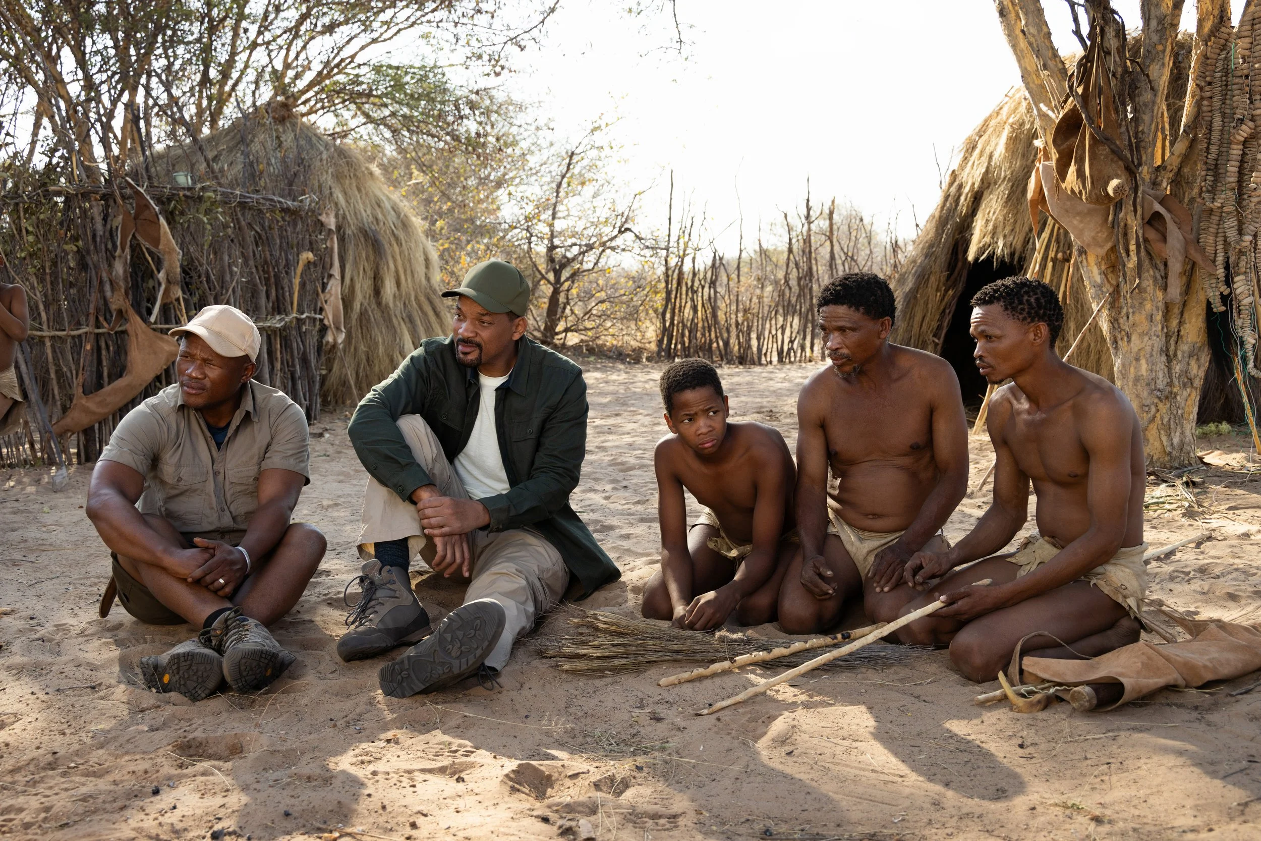 Will smith in a group of three men and a boy sitting on the ground in front of traditional huts made of sticks and grass, in the Kalahari Desert with leafless trees in the background.
