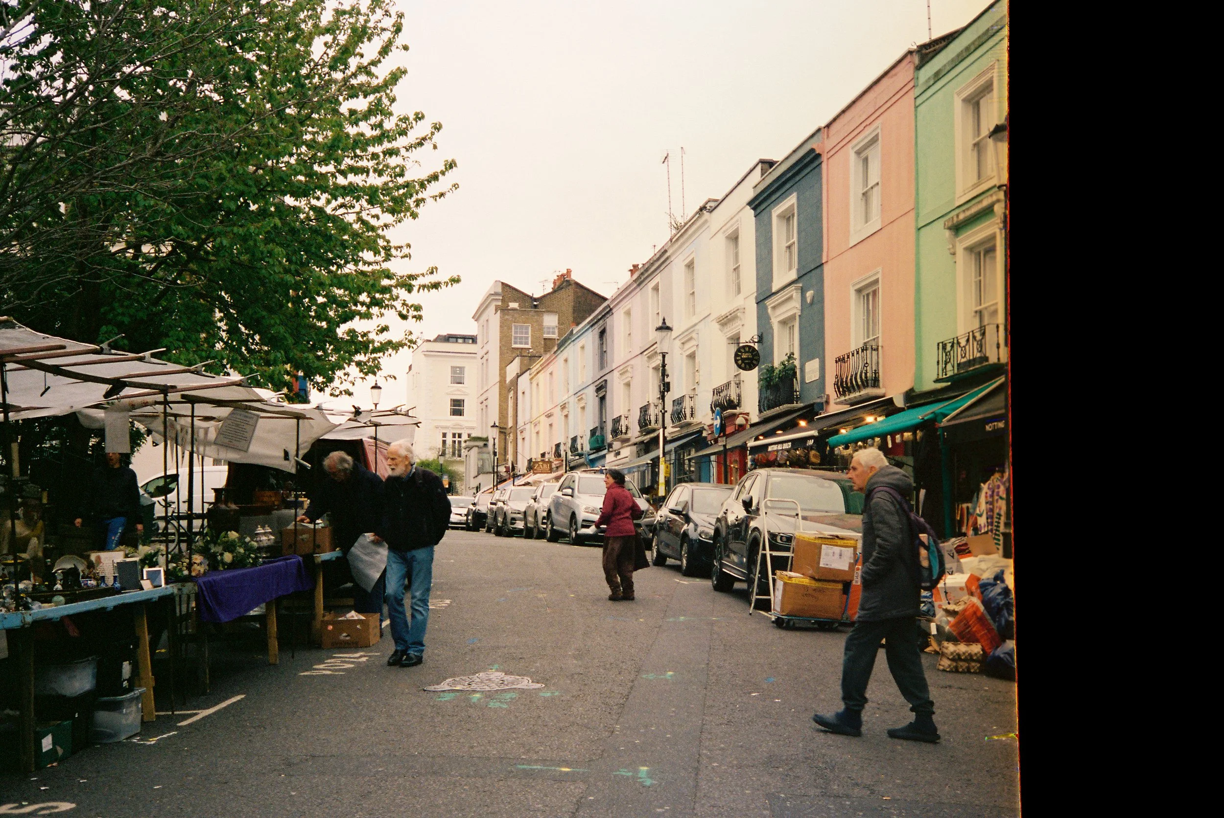 Street market scene with colorful row houses, outdoor vendors, and people shopping on a cloudy day.  Photographed by Julia Tran in London, United Kingdom. Photographed on 35mm film. 