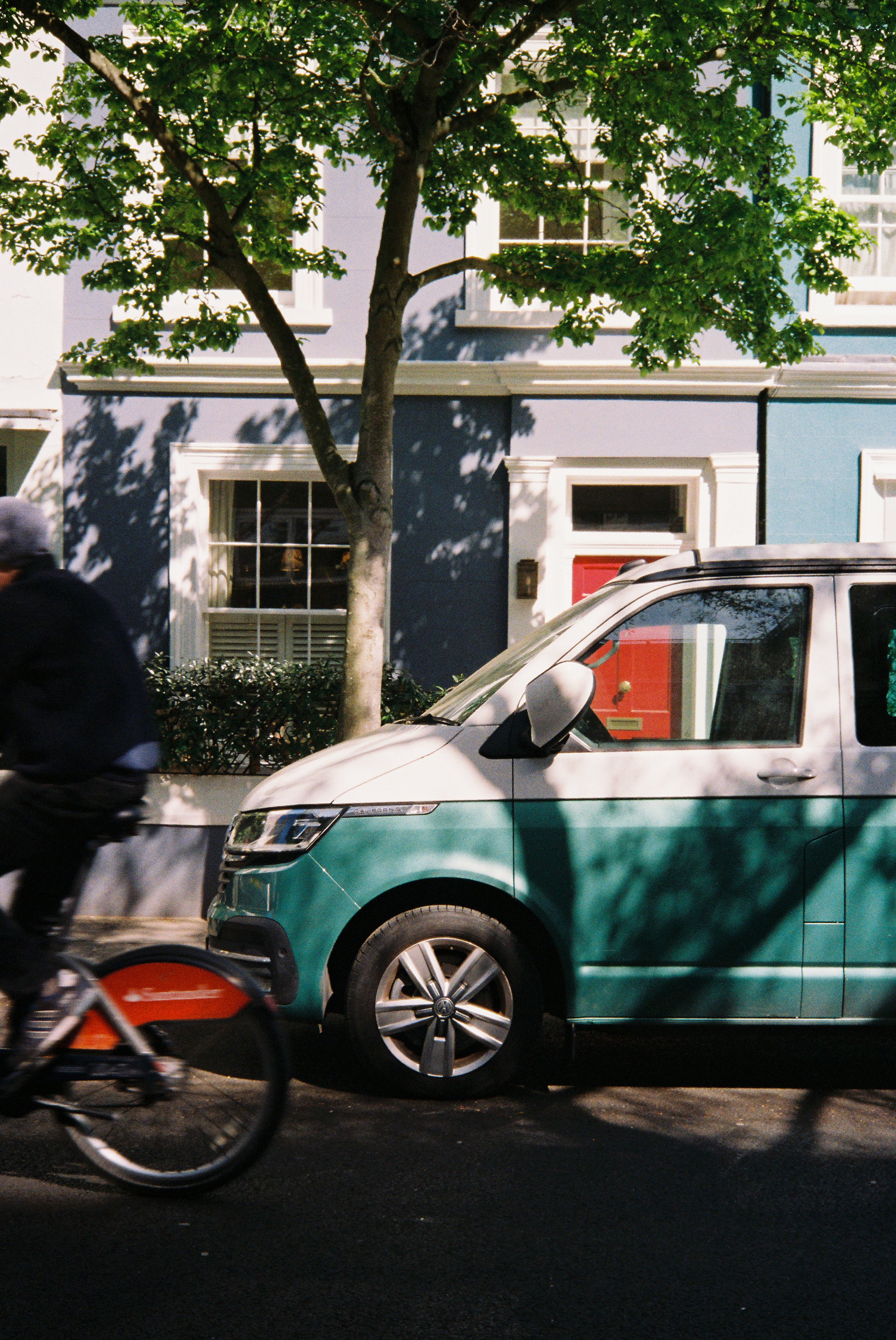 A street with a green and white vehicle passing by, a tree casting shadows on the buildings, and a person riding a bicycle in motion in the foreground.  Photographed by Julia Tran in London, United Kingdom. Photographed on 35mm film. 
