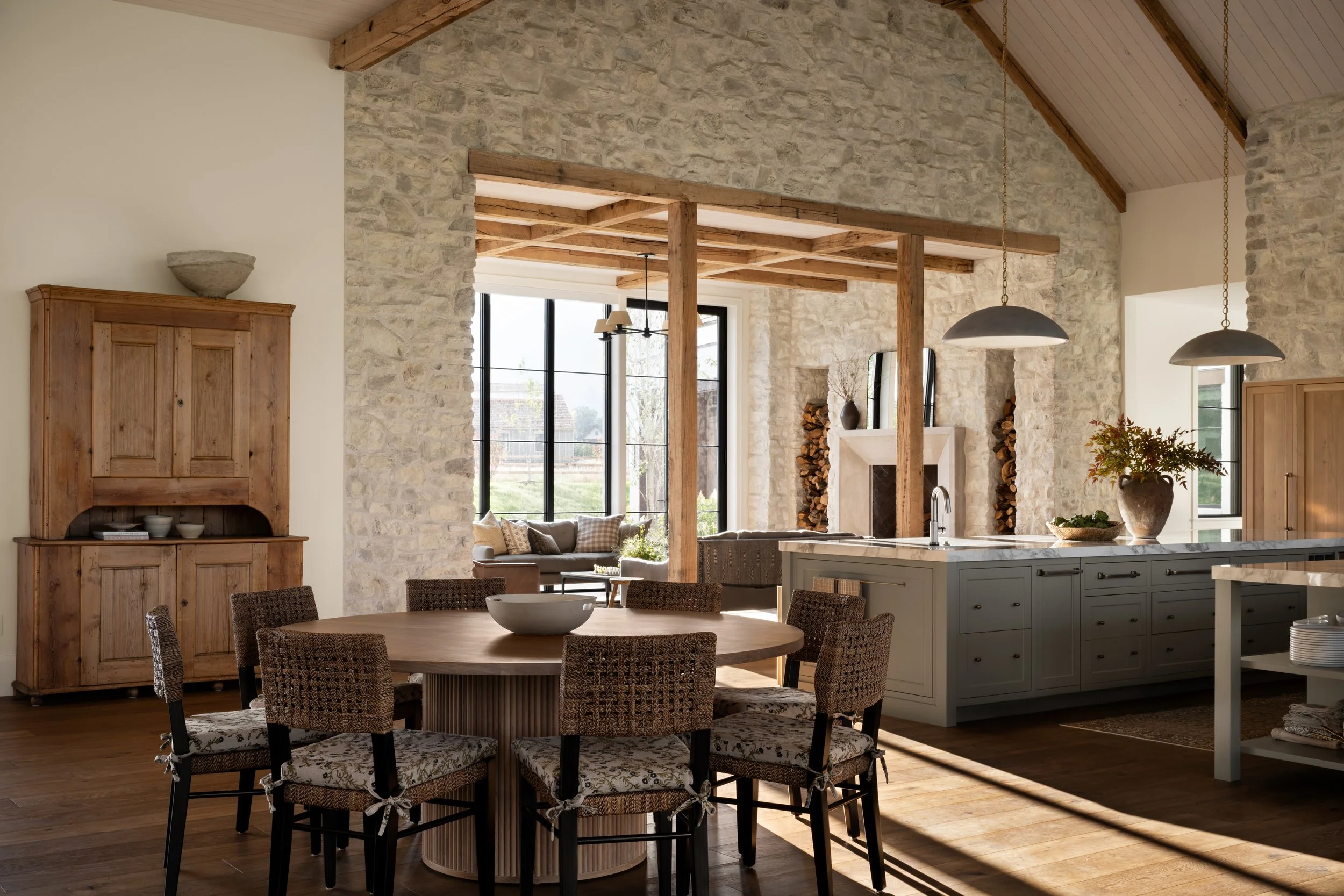 In a space designed by Studio McGee is a living room and kitchen with stone walls, wooden beams, large windows, and a shaded outdoor view. There is a round dining table with wicker chairs, and a kitchen island with a decorative bowl and plant.
