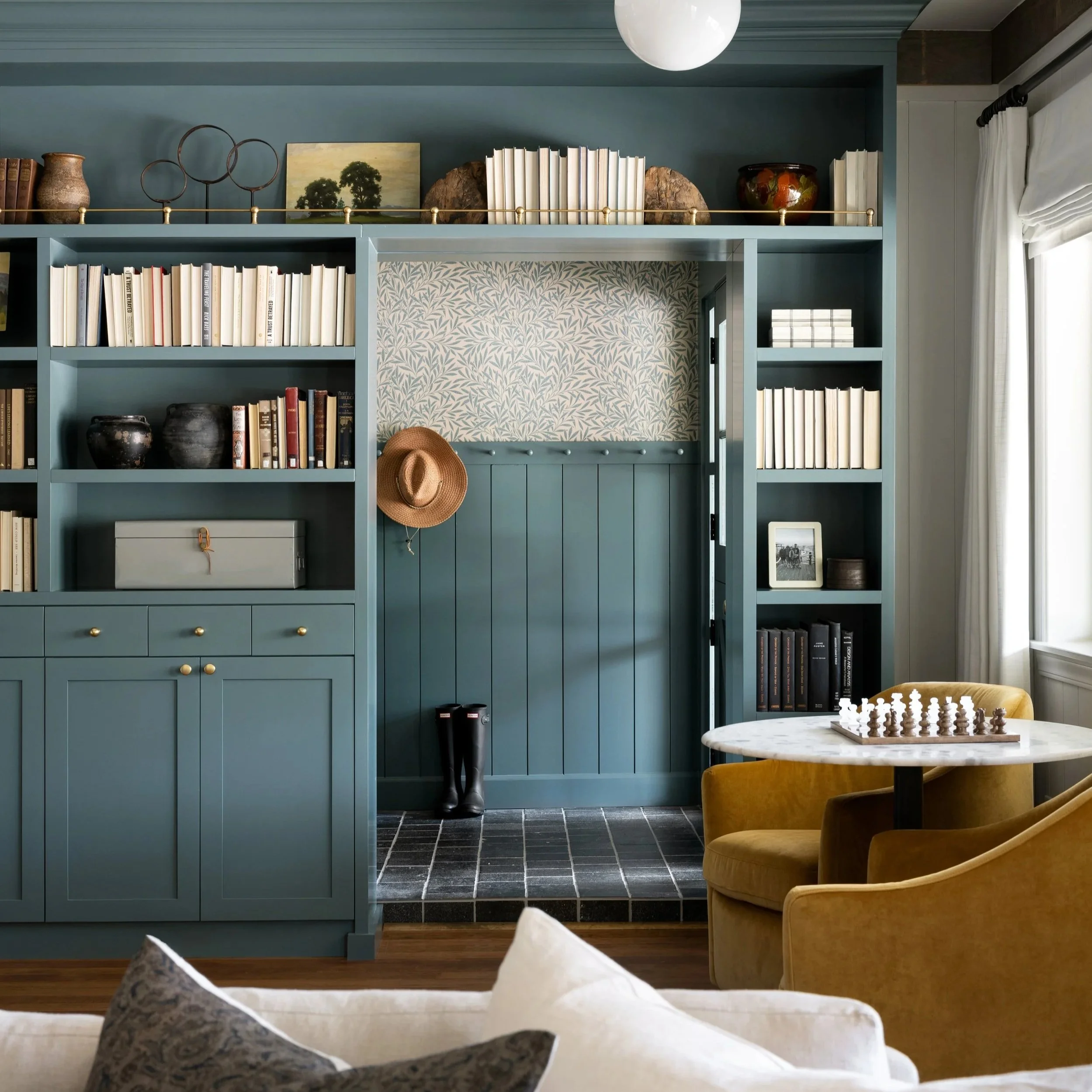 A cozy living room designed by Studio McGee with a built-in blue bookshelf filled with books, decorative vases, and artwork. A round marble table with a chess set and a yellow armchair are visible.