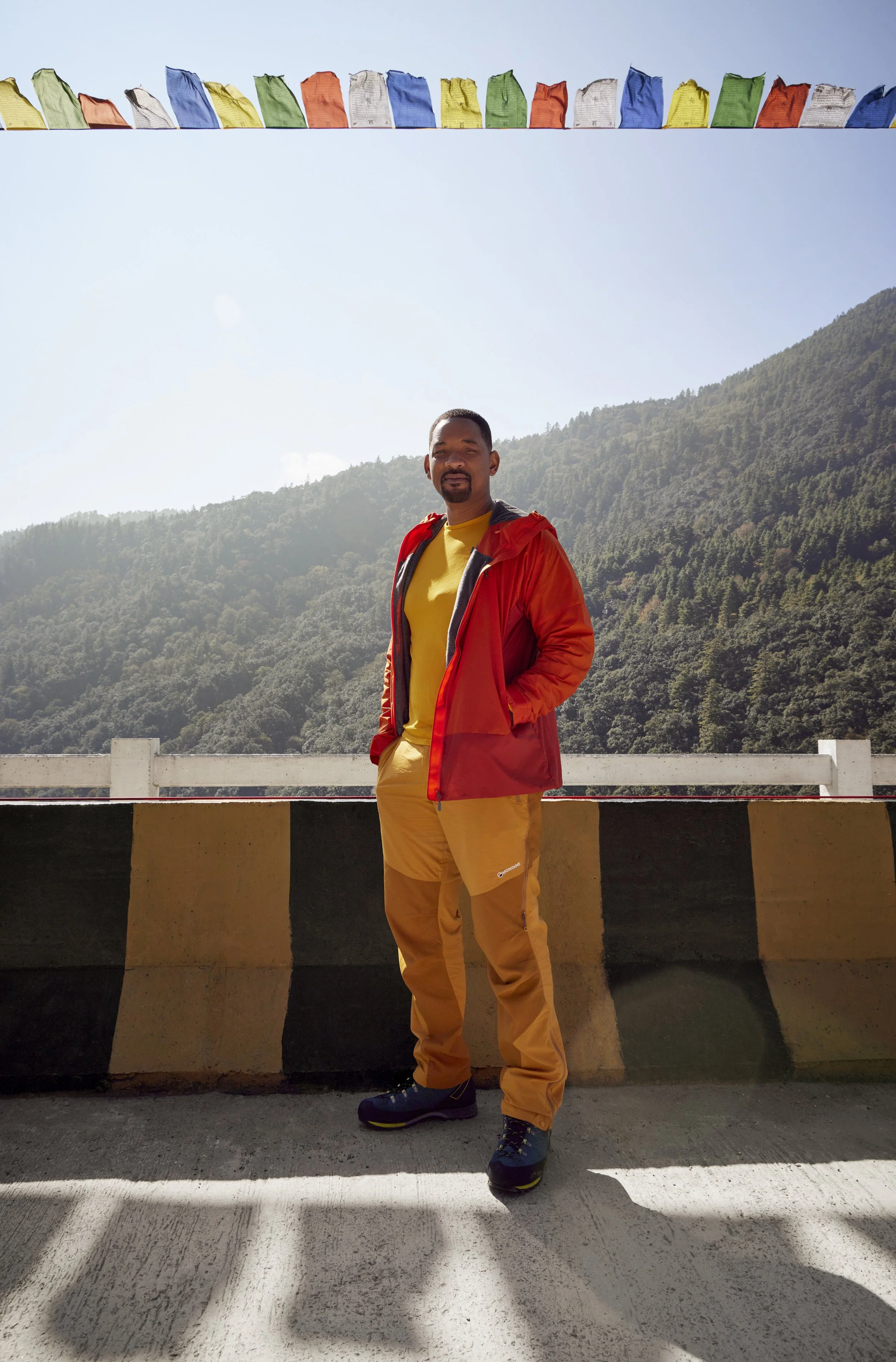 Will Smith in outdoor hiking gear standing outdoors against a mountainous background, with prayer flags hanging above him.