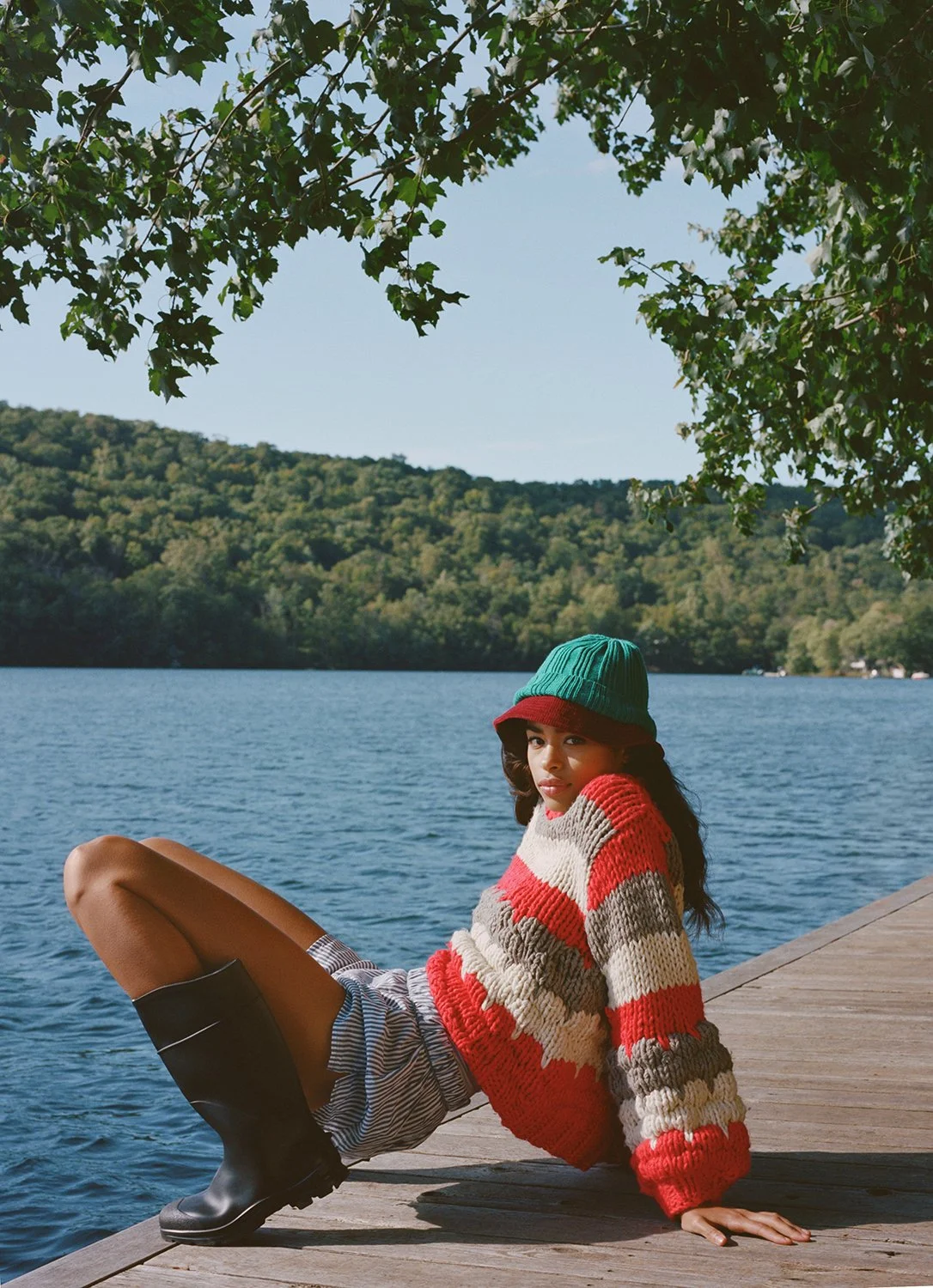 A woman wearing a green hat, red and gray striped sweater, striped shorts, and black rubber boots sitting on a wooden dock by a lake, with green trees and hills in the background for Urban Outfitters.