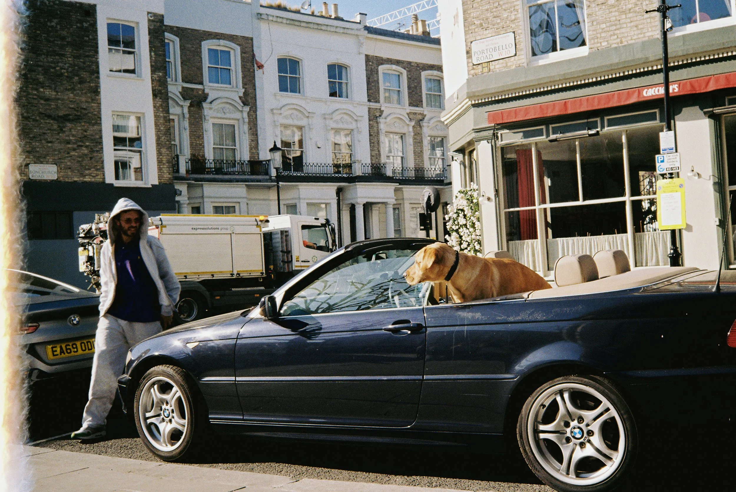 A man in a white hoodie and gray pants walking past a black BMW convertible with a dog in the driver's seat, parked on a city street with buildings and vehicles in the background.  Photographed by Julia Tran in London, United Kingdom. 35mm film.