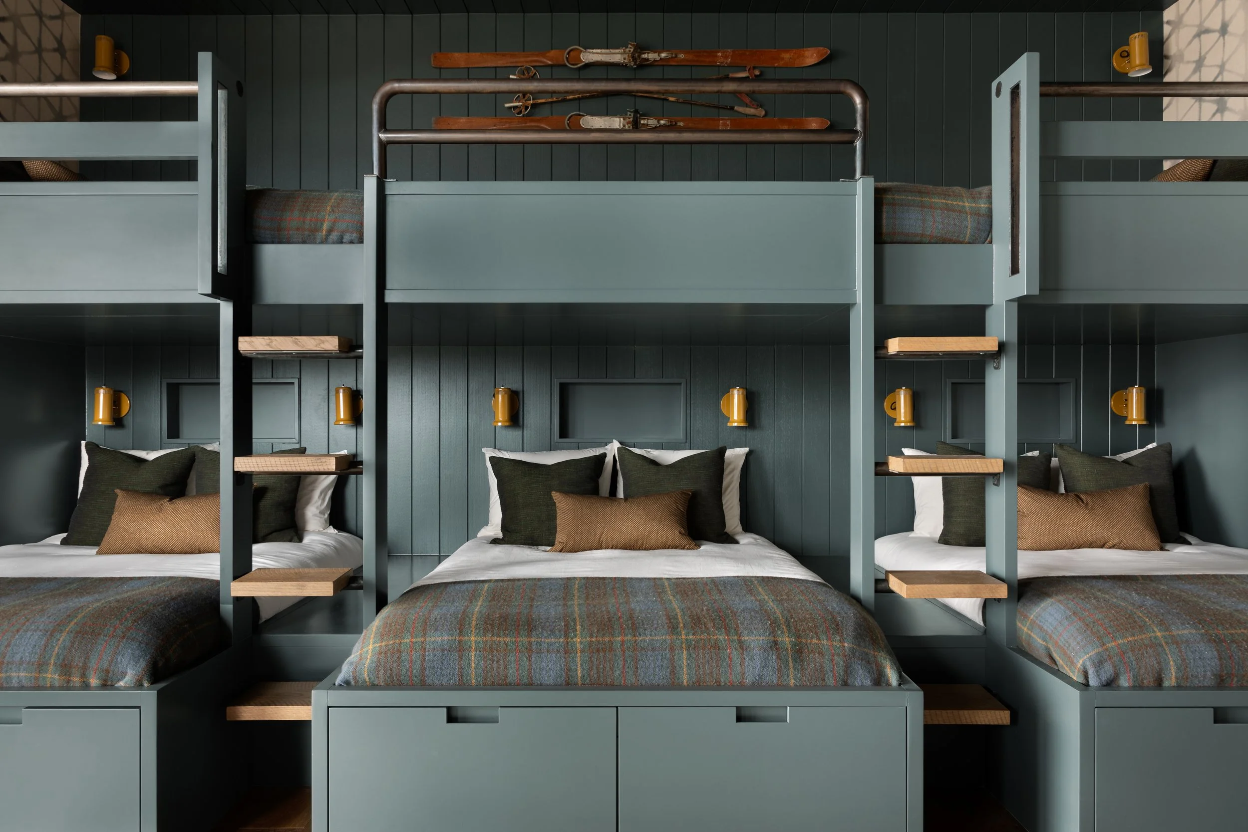 In a space designed by Studio McGee, a wall with three brown skis mounted horizontally, above a bed with multiple pillows and a dark green headboard, featuring built-in shelves and yellow wall-mounted lamps.
