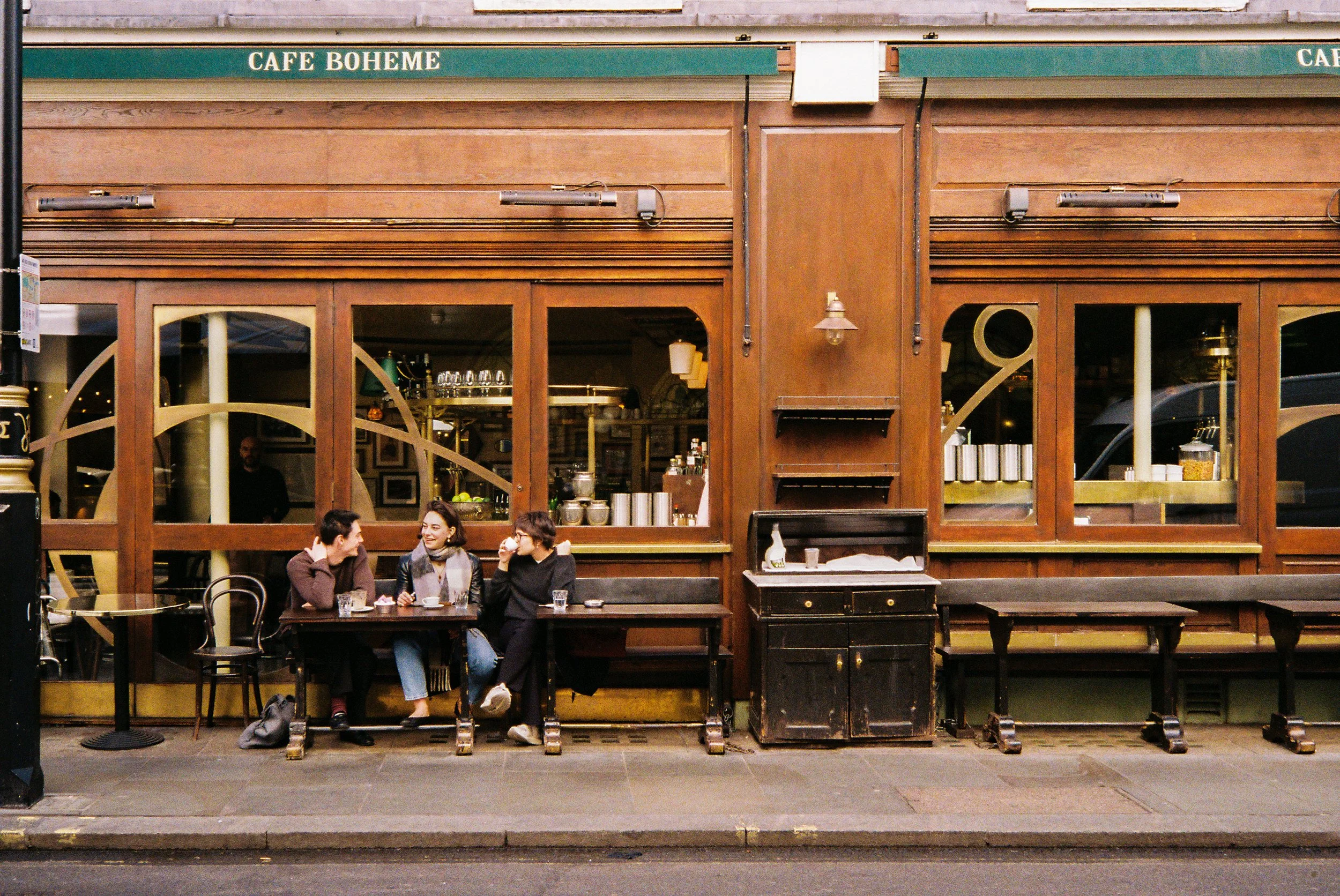 A wooden cafe with a green sign reading 'Café Boheme'. Outside, four people sit at a table chatting, with drinks on the table. The cafe has large windows and a door, with reflections of the street and a parked car visible.  Photographed by Julia Tran