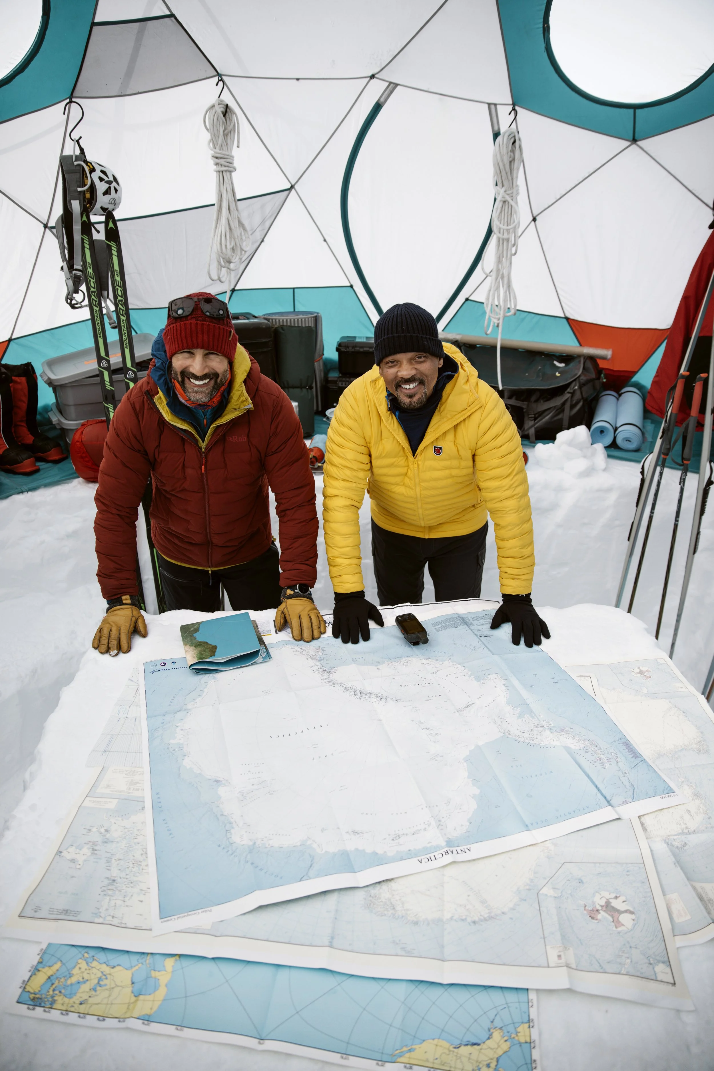 Will Smith & Friend in winter gear inside a snow-covered tent, standing behind a large map on a table, smiling at the camera.