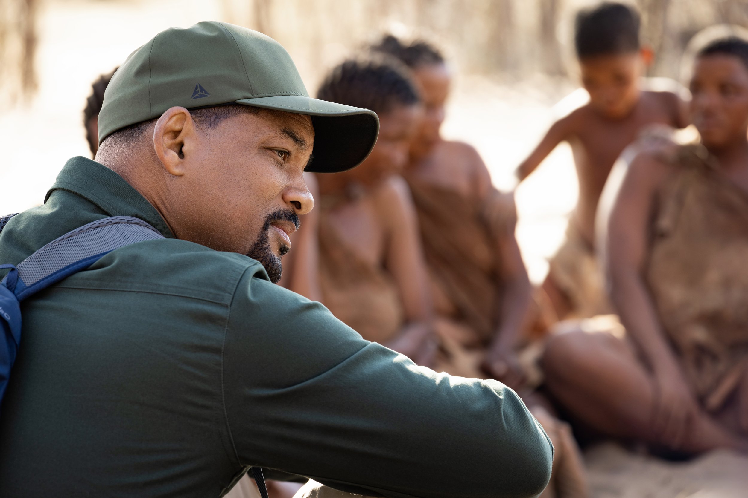 Will Smith in outdoor clothing and a cap sitting and listening, with several children sitting in the background.