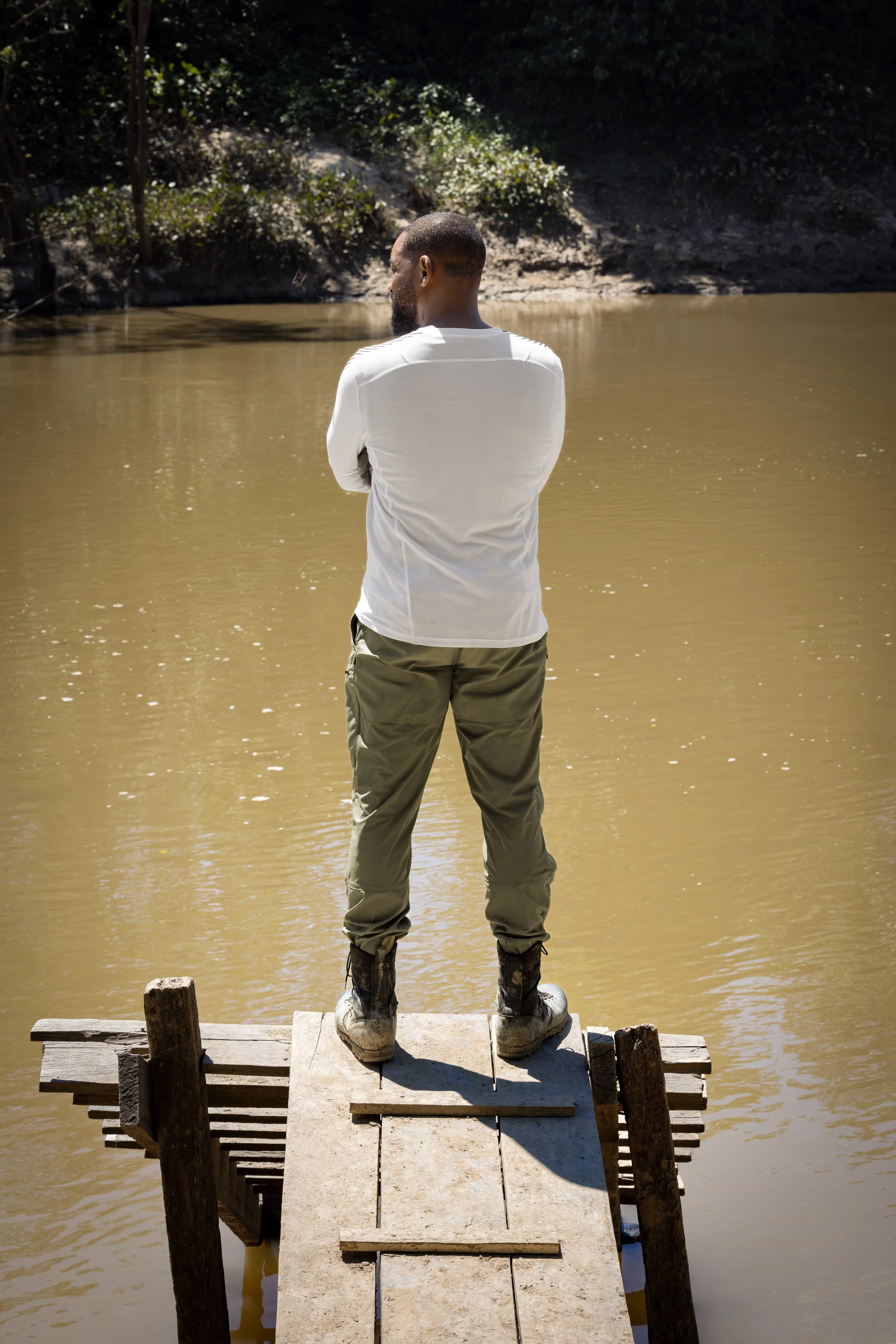 Will Smith on a small wooden dock, facing away towards a muddy pond or lake surrounded by trees and bushes, with his arms crossed.