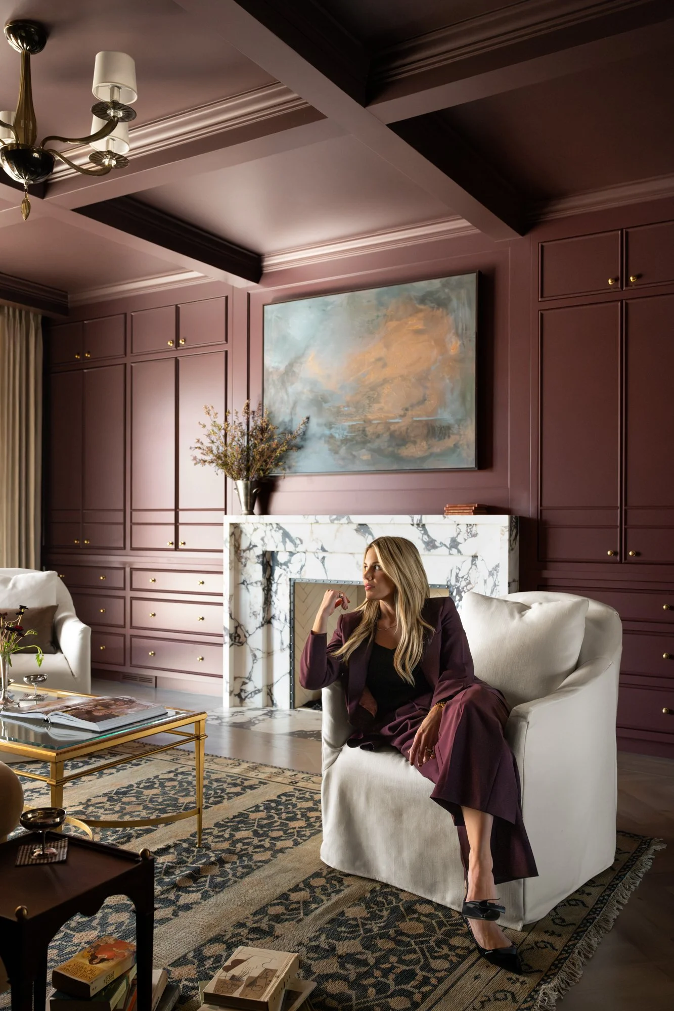 Shea McGee sitting on a white armchair in a luxurious living room with purple paneled walls, a marble fireplace, a large abstract painting, and a gold chandelier. She is wearing a maroon suit and black high heels.