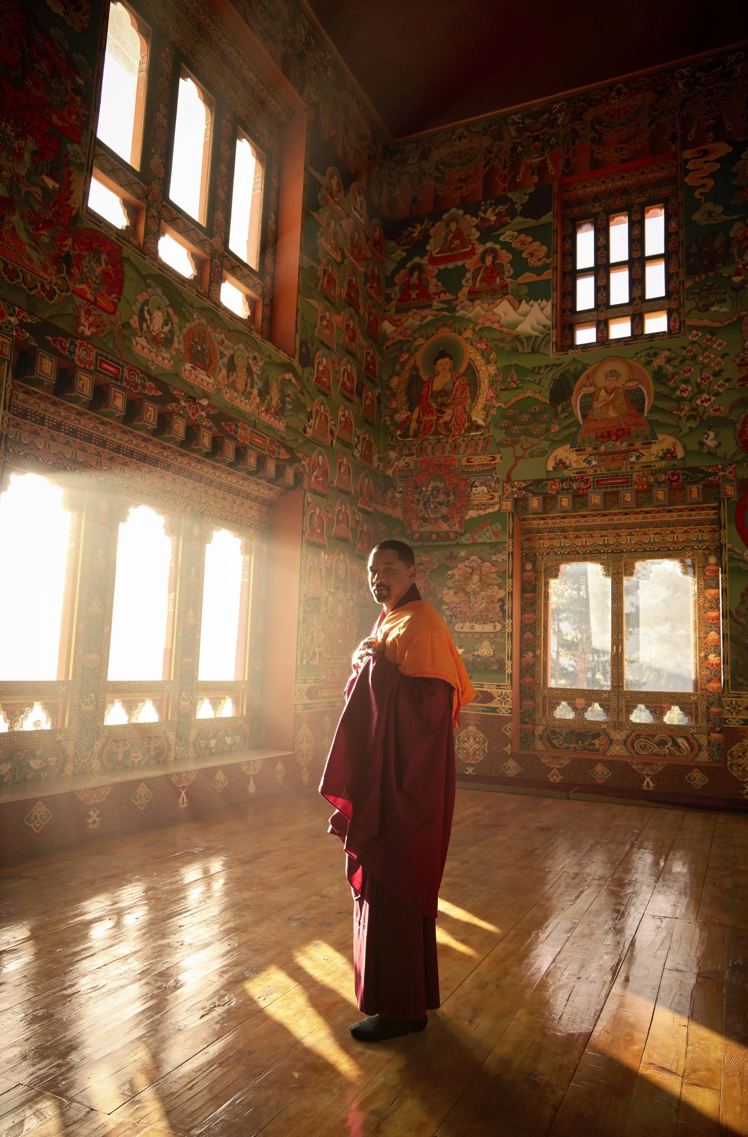 Will Smith standing inside a brightly colored temple, illuminated by natural sunlight coming through the windows, with intricate wall murals. 