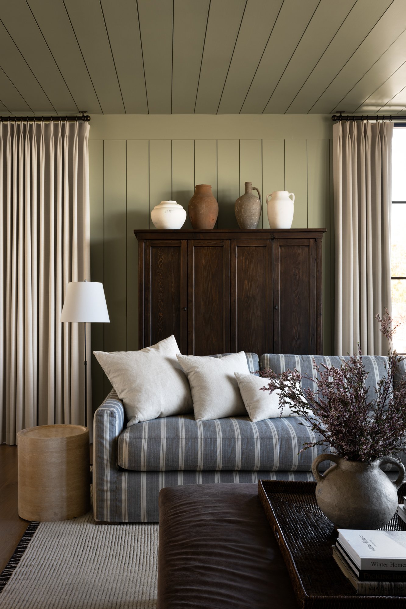 Designed by Studio McGee, is a living room with green walls, dark wood cabinet, white curtains, beige pillows on blue striped sofa, white floor lamp, vase with purple flowers, and a tray with books on a dark brown ottoman.