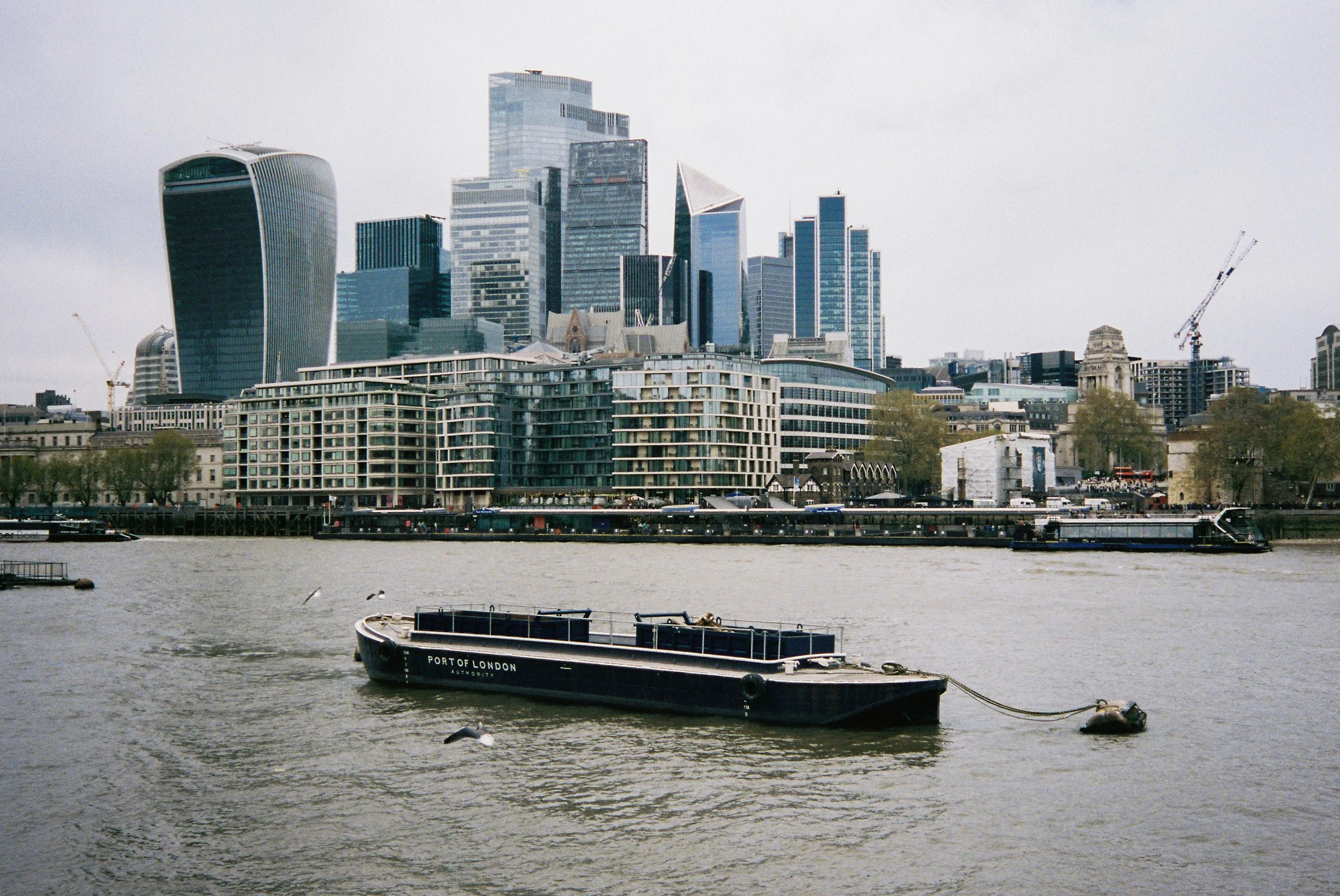 View of a city skyline with modern skyscrapers and buildings along the riverbank, including a boat labeled 'Port of London' in the foreground on the water.  Photographed by Julia Tran in London, United Kingdom. Photographed on 35mm film. 