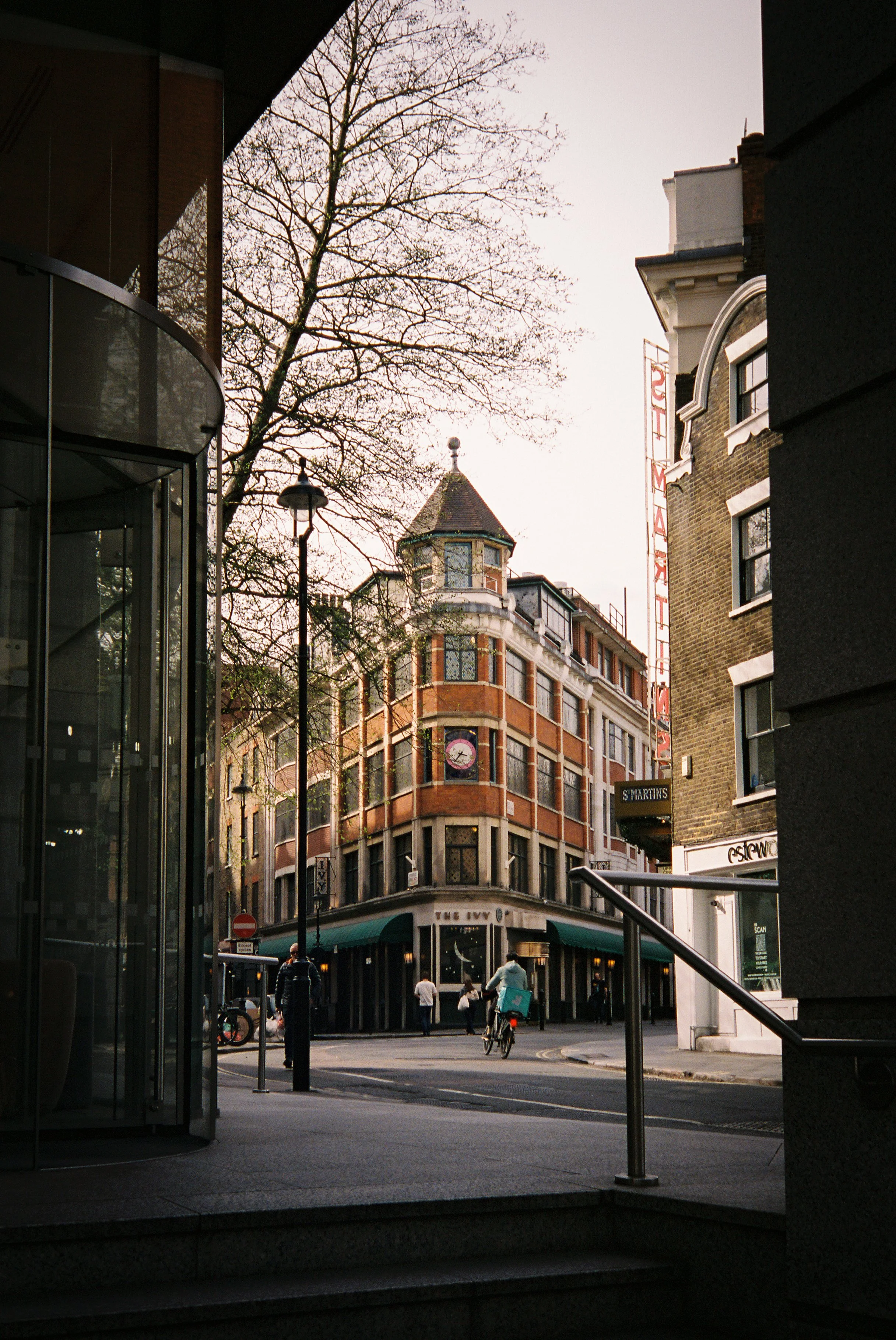 A city street scene viewed from a building entrance, with a mix of old and new architecture, pedestrians, cyclists, and street signs during daytime, under a cloudy sky.  Photographed by Julia Tran in London, United Kingdom. Photographed on 35mm film.