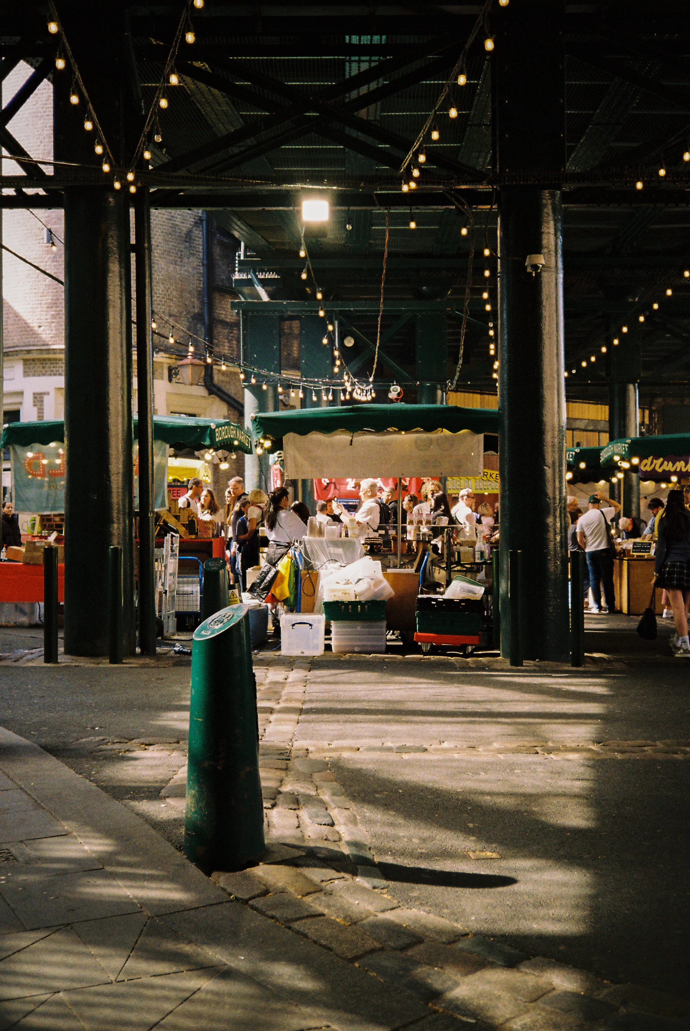 Market stall set up under a bridge with string lights, people shopping and vendors in the background, daytime outdoor scene.  Photographed by Julia Tran in London, United Kingdom. Photographed on 35mm film. 