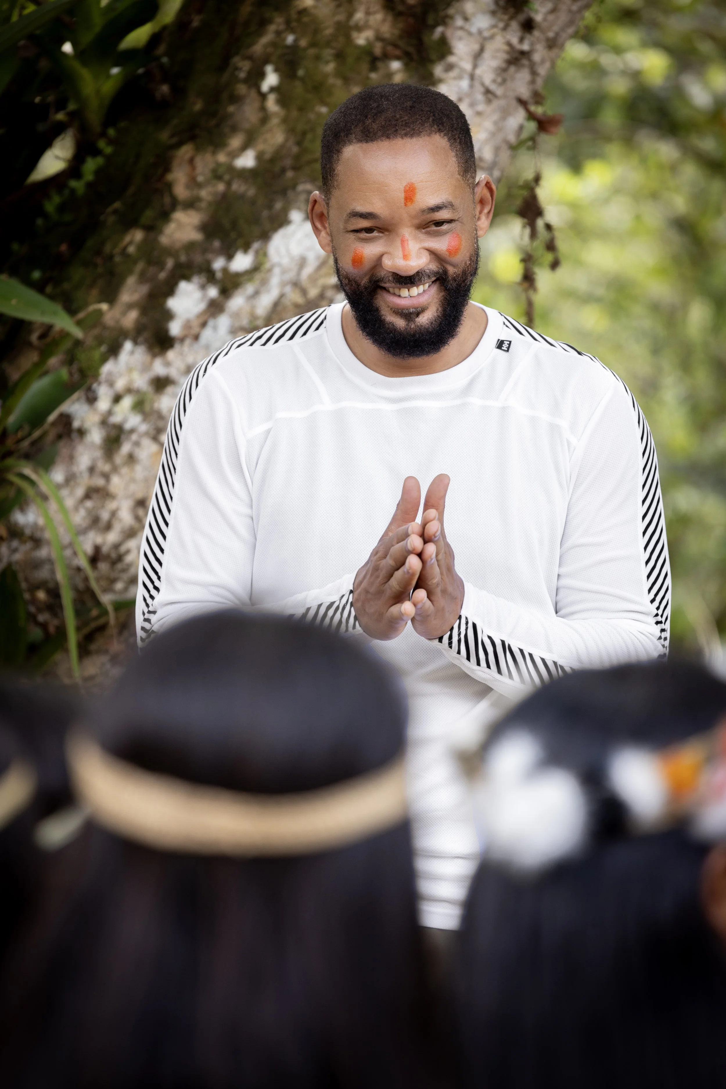Will Smith smiling, dressed in white with red face paint, standing outdoors against a tree, engaging with a crowd.