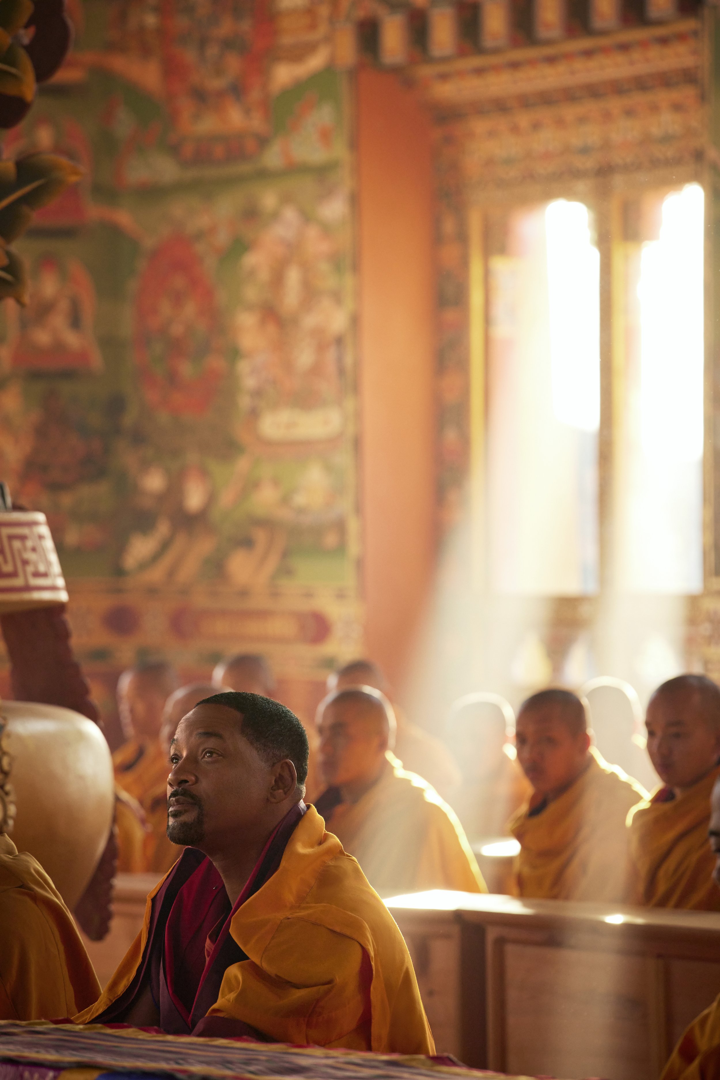 Will Smith & Monks sitting in prayer inside a brightly lit temple with colorful wall murals.